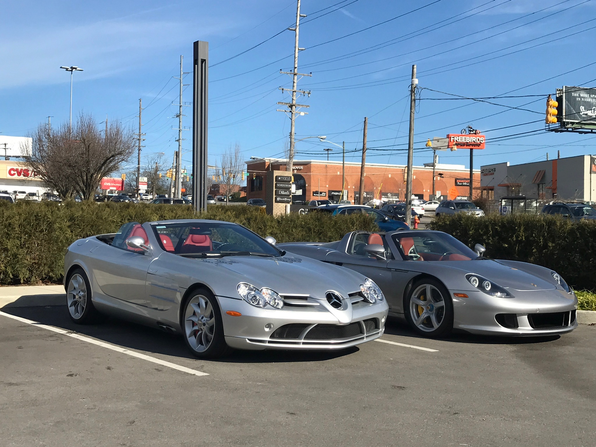 Two silver convertible sports cars are parked side by side in a parking lot on a sunny day, with shops and power lines visible in the background.
