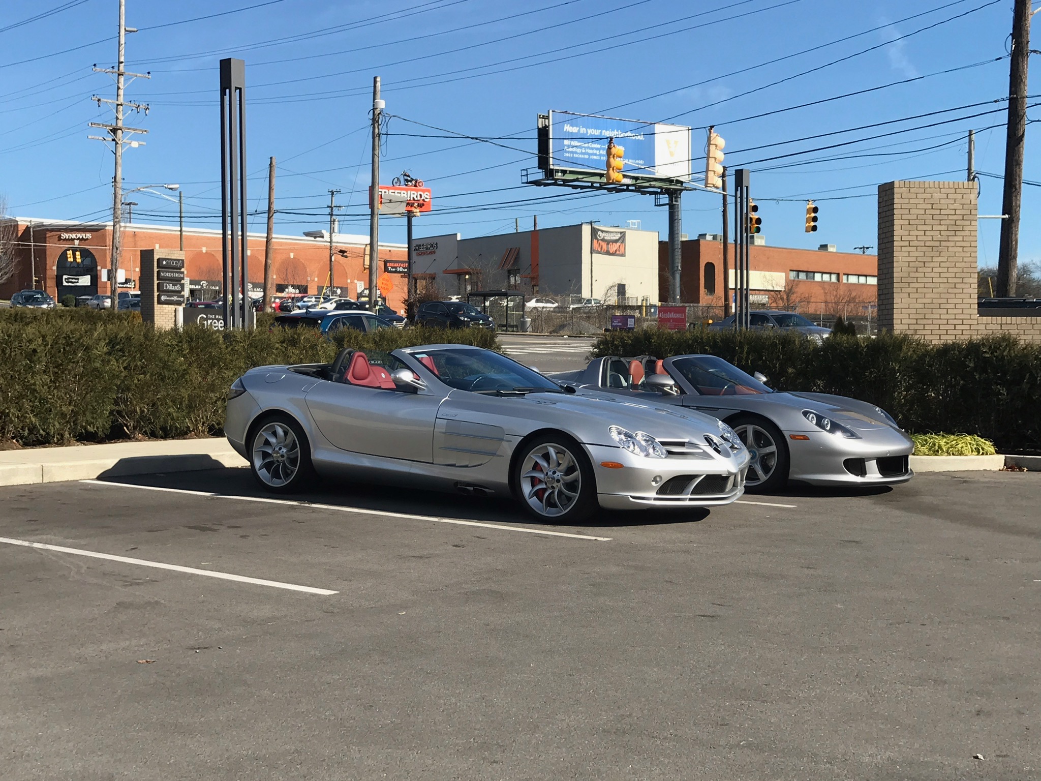 Two silver convertible sports cars are parked side by side in a nearly empty parking lot on a sunny day, with commercial buildings and power lines in the background.