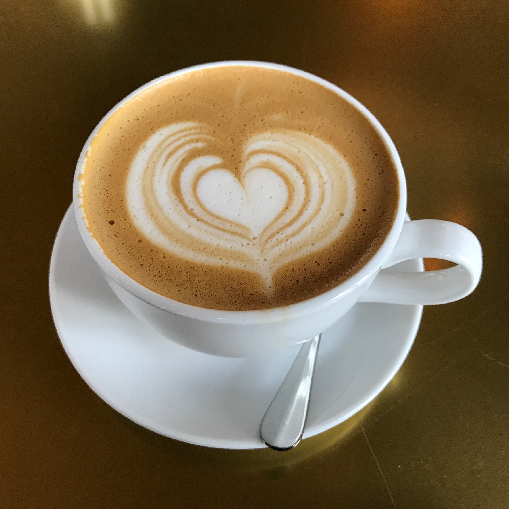 A cup of coffee with heart-shaped latte art sits on a white saucer with a metal spoon, against a gold background.