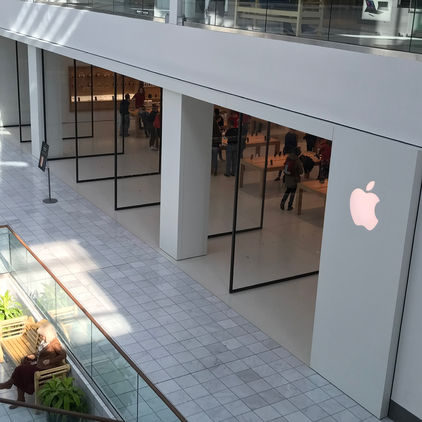 Apple Store in a modern indoor shopping mall, with a large white Apple logo on the storefront and people inside browsing products.