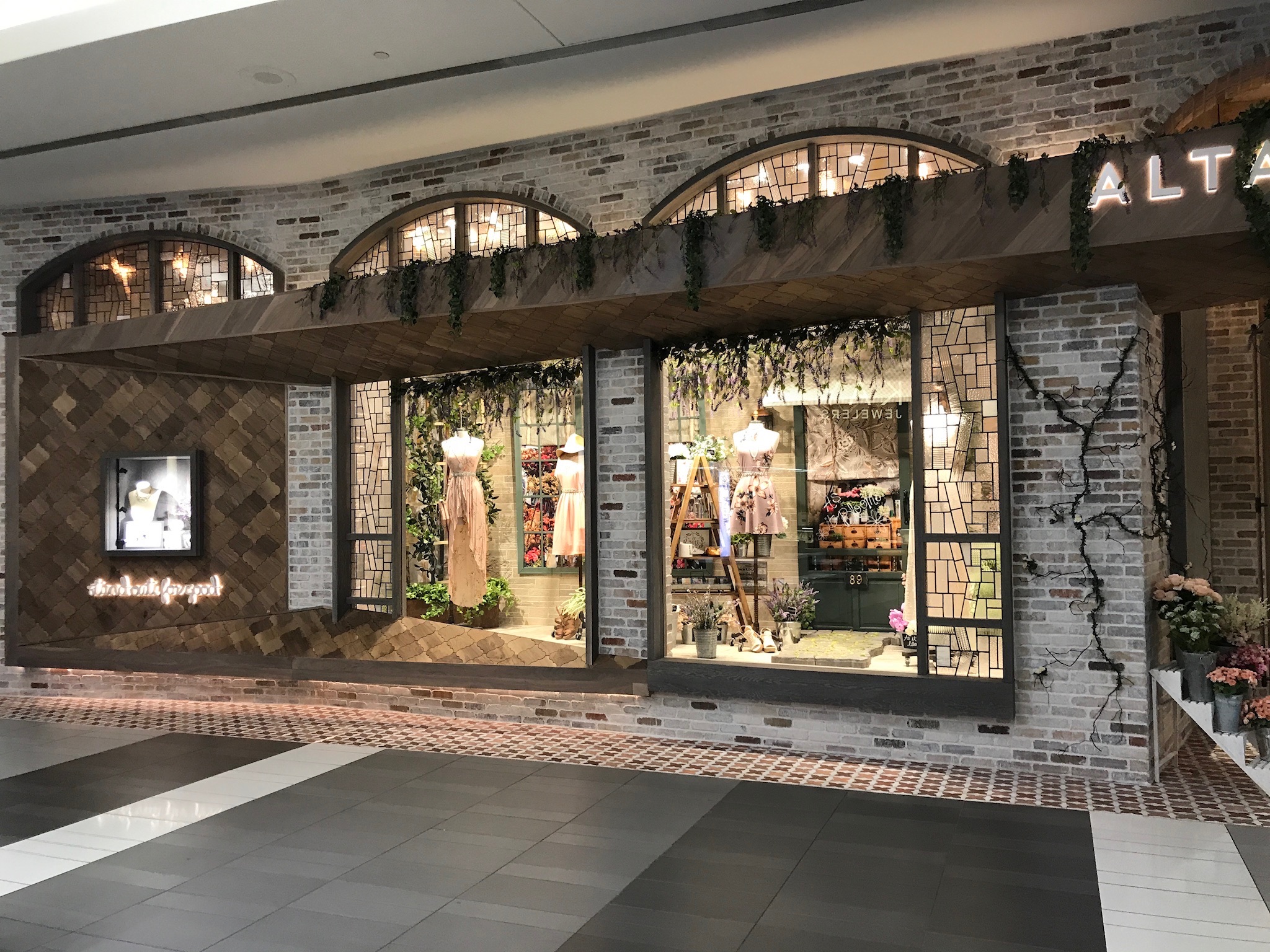 Shopfront with a large display window featuring clothing and accessories, brick and wood decor, and plants hanging above the window.