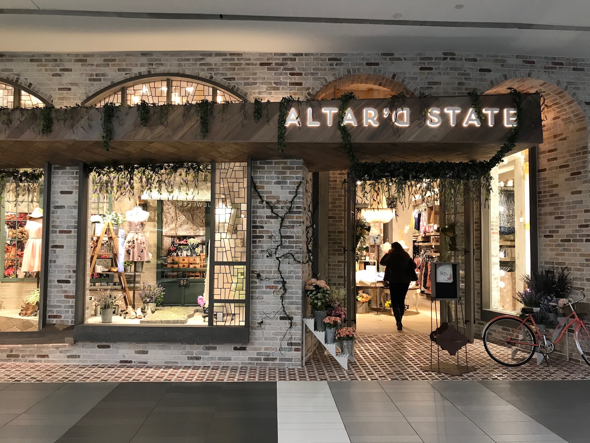 A storefront for Altar'd State in a mall, featuring a brick facade, window displays with clothes, and a person entering through the main door. A bicycle is parked outside.
