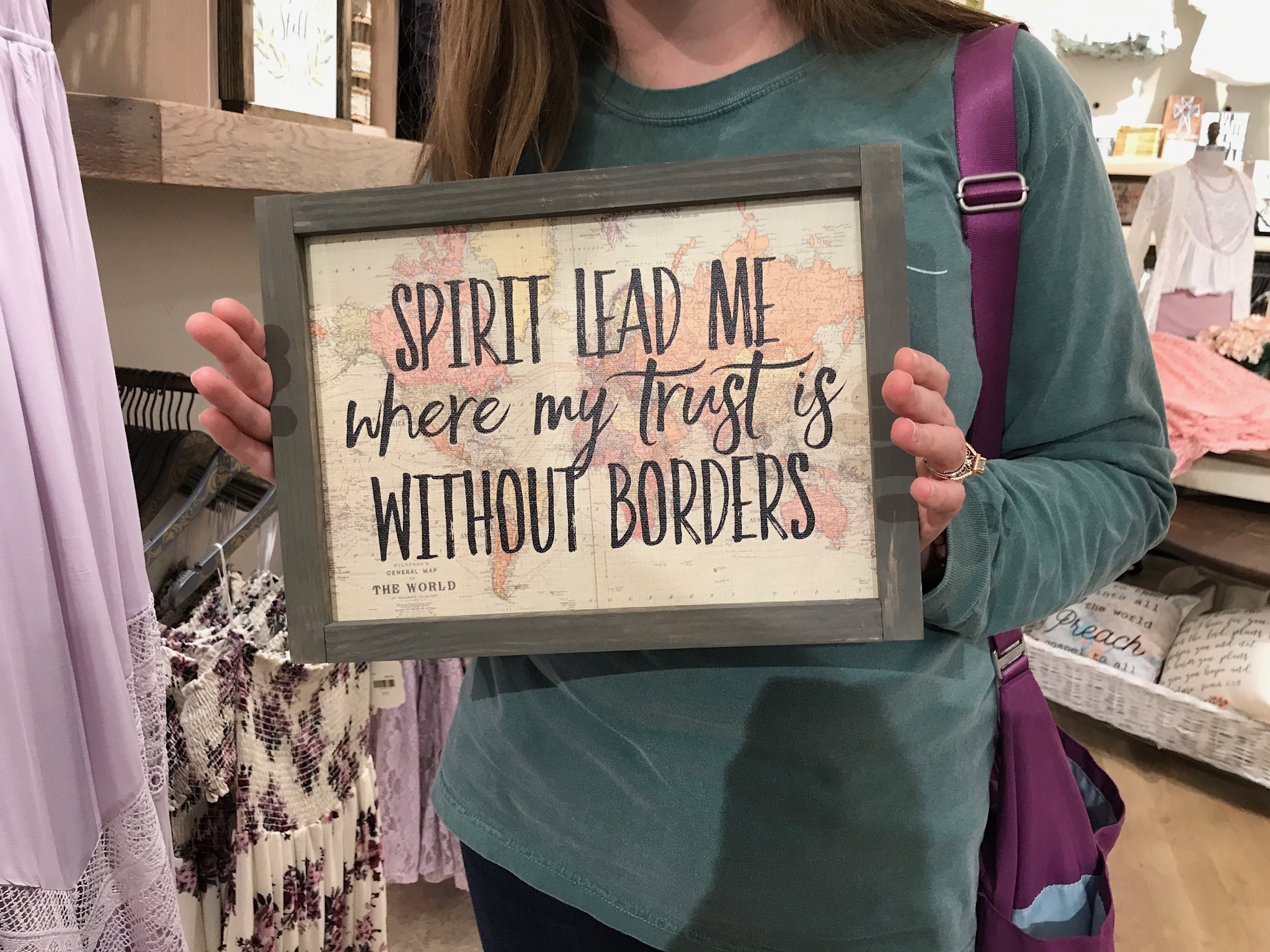 Person holding a framed sign that reads, "Spirit lead me where my trust is without borders," in a store with clothing and decor items in the background.