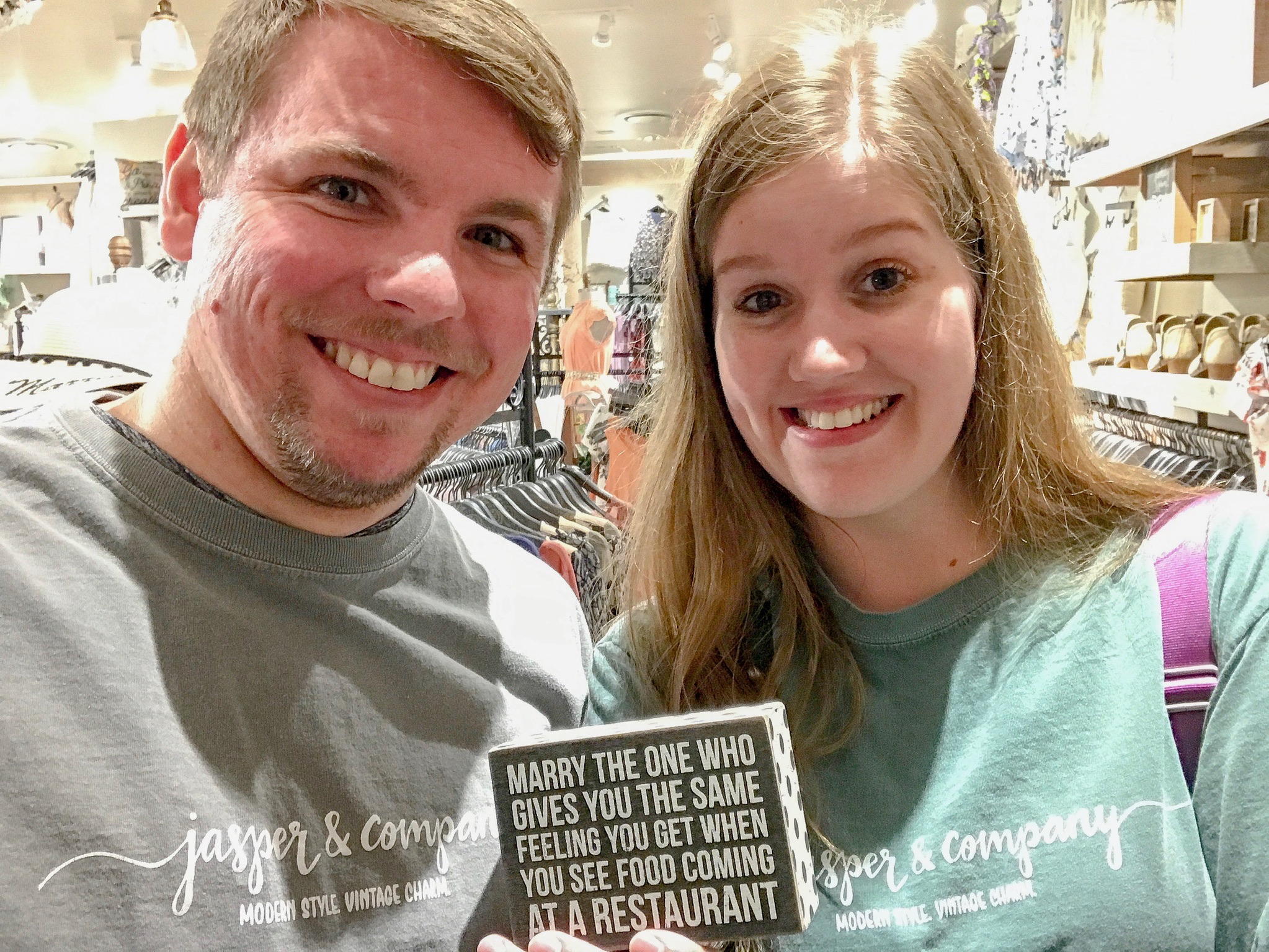 Two people smile at the camera in a store, both wearing "jasper & company" shirts. The woman holds a sign that says, "Marry the one who gives you the same feeling you get when you see food coming at a restaurant.