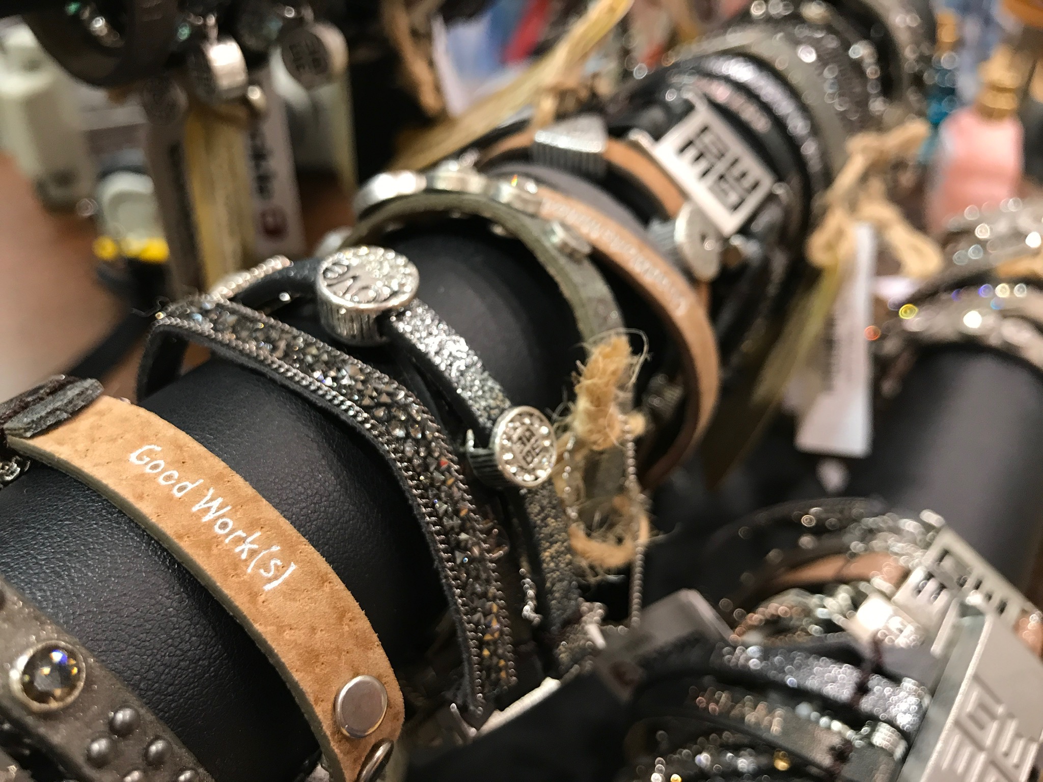 Close-up of several beaded, leather, and metal bracelets displayed on a black stand, including one labeled "Good Work(s).