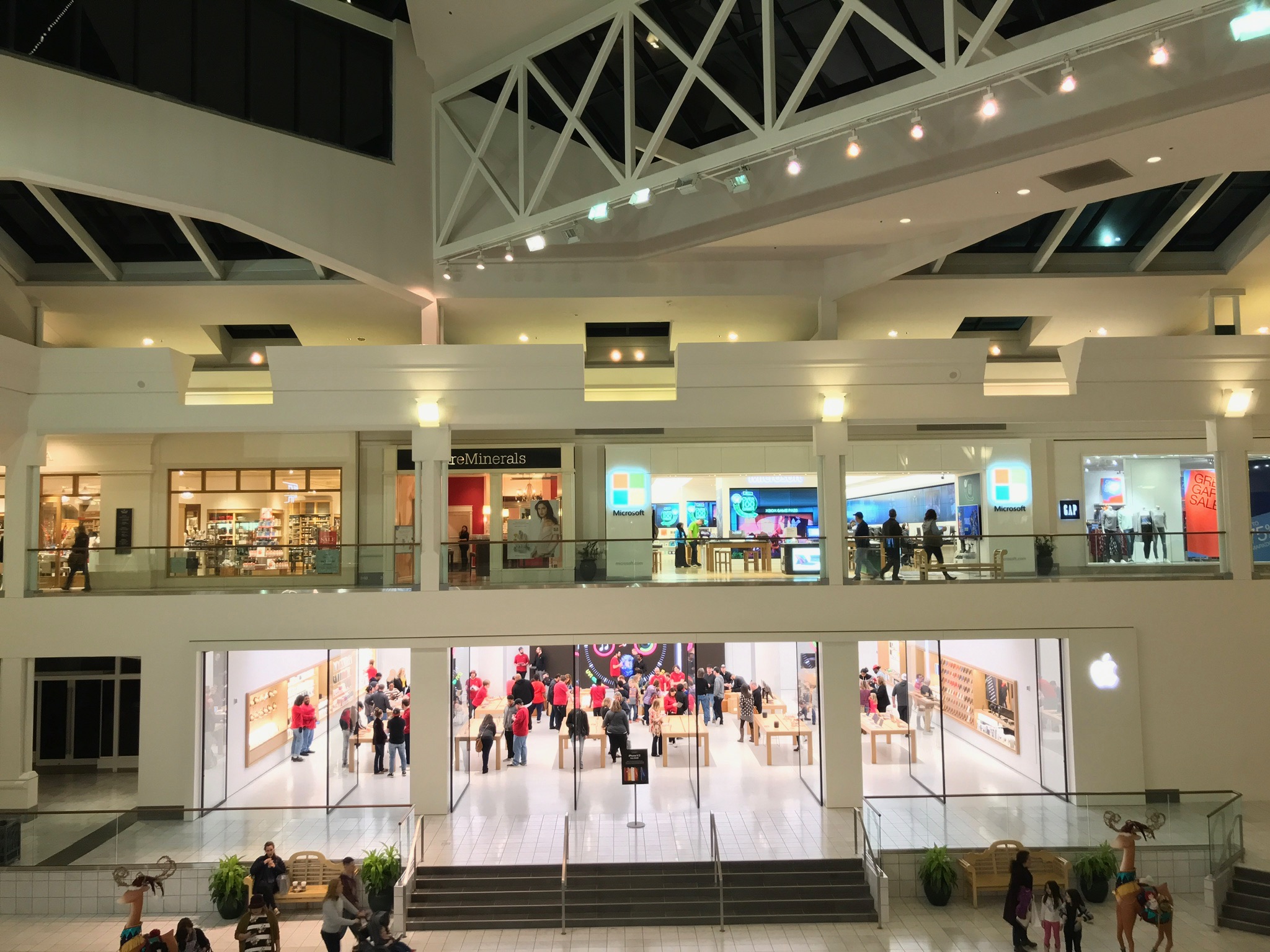 A shopping mall interior with two floors, featuring stores including an Apple Store on the lower level and a Microsoft Store on the upper level. Shoppers are visible throughout.