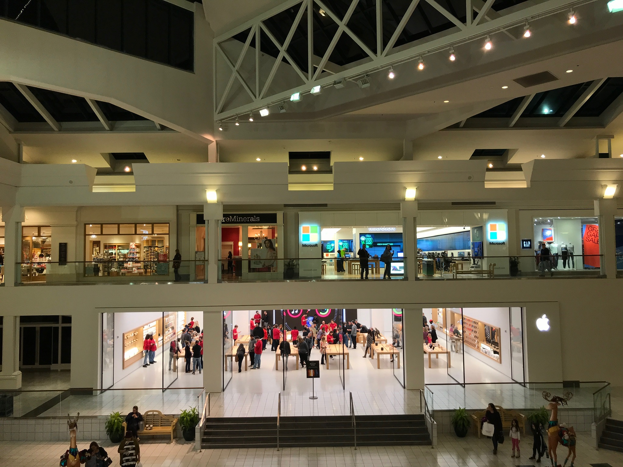 Interior view of a shopping mall showing two levels with various stores, including an Apple Store on the lower level and a Microsoft Store on the upper level. Shoppers are visible throughout.