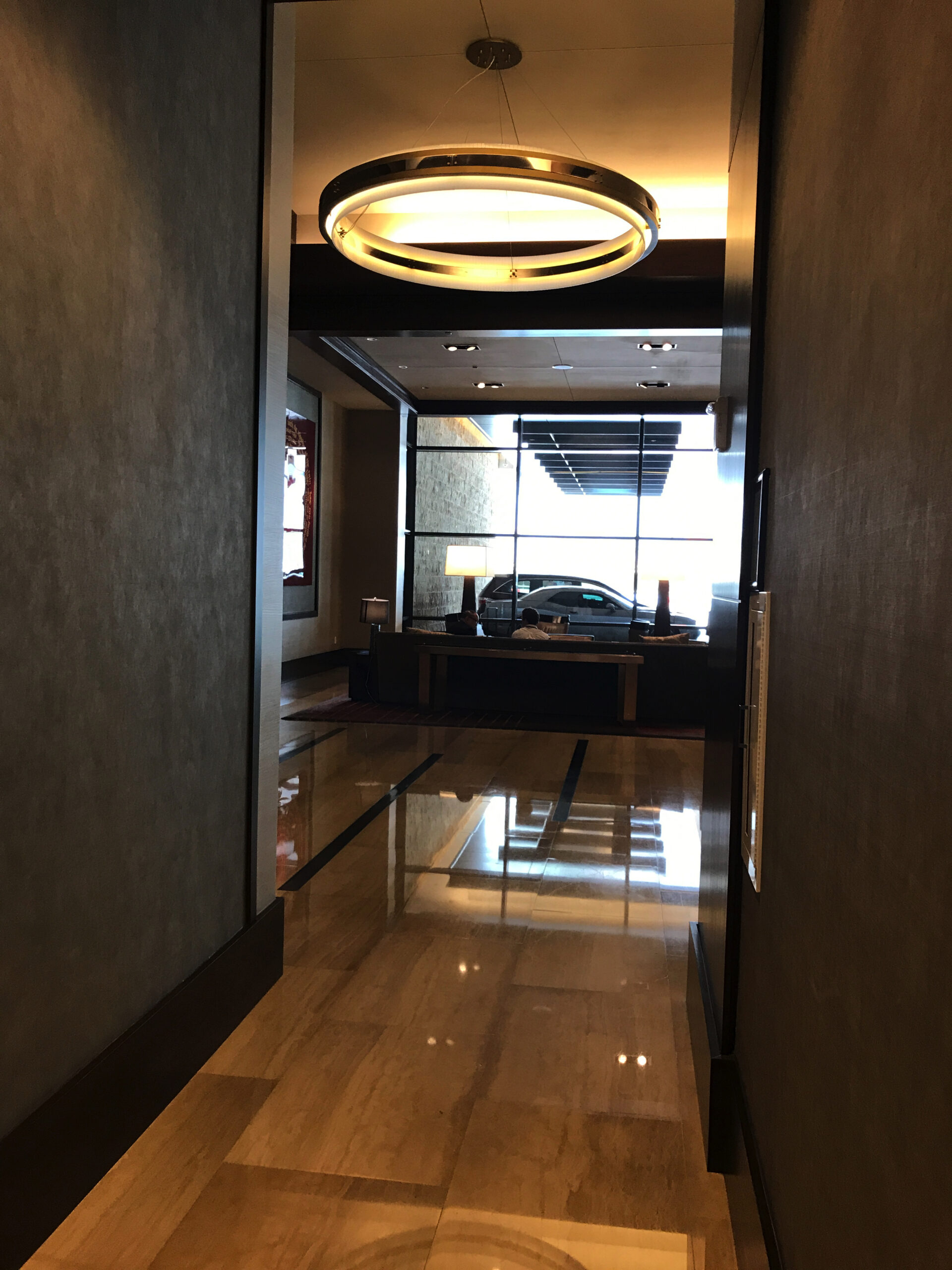 A hallway with polished marble floors leads to a windowed lobby area where a car is parked outside; a circular ceiling light hangs above.