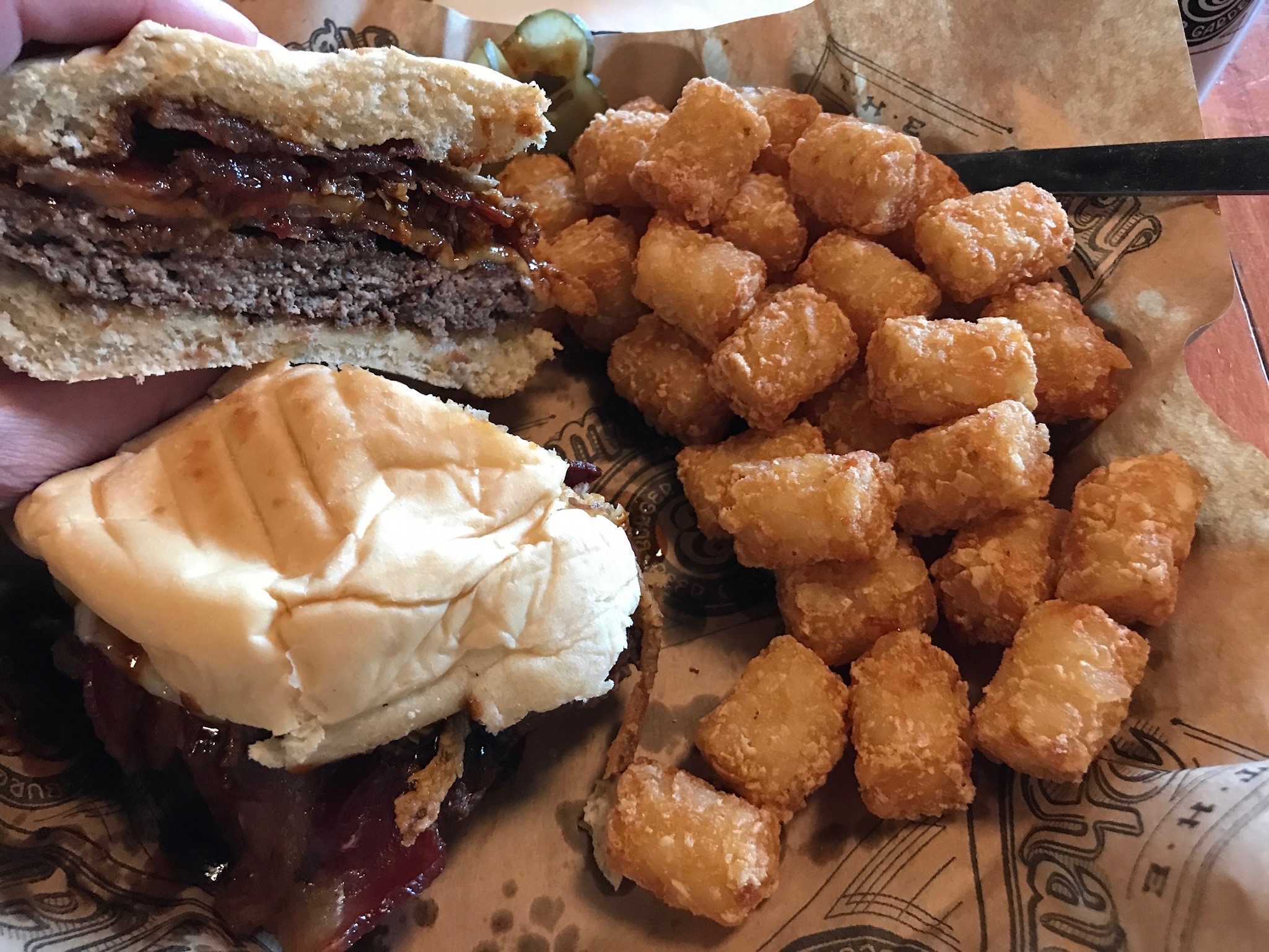 A cheeseburger with a bite taken out is held above a plate of golden tater tots and another half of the sandwich on brown paper.