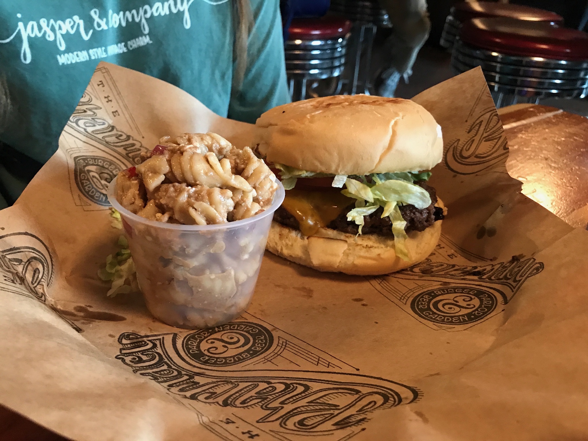 A cheeseburger with lettuce and tomato sits on branded paper next to a small plastic cup of pasta salad on a wooden table.