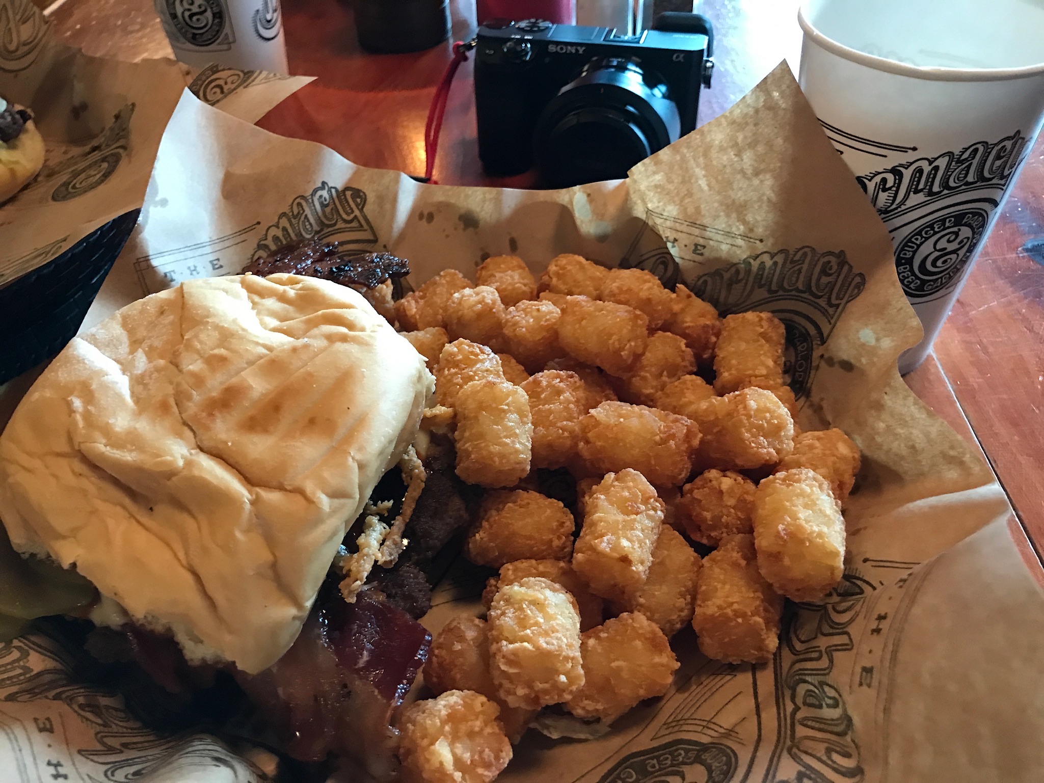 A cheeseburger with bacon and crispy onion, served with tater tots on brown wax paper, with a drink cup and a camera in the background.