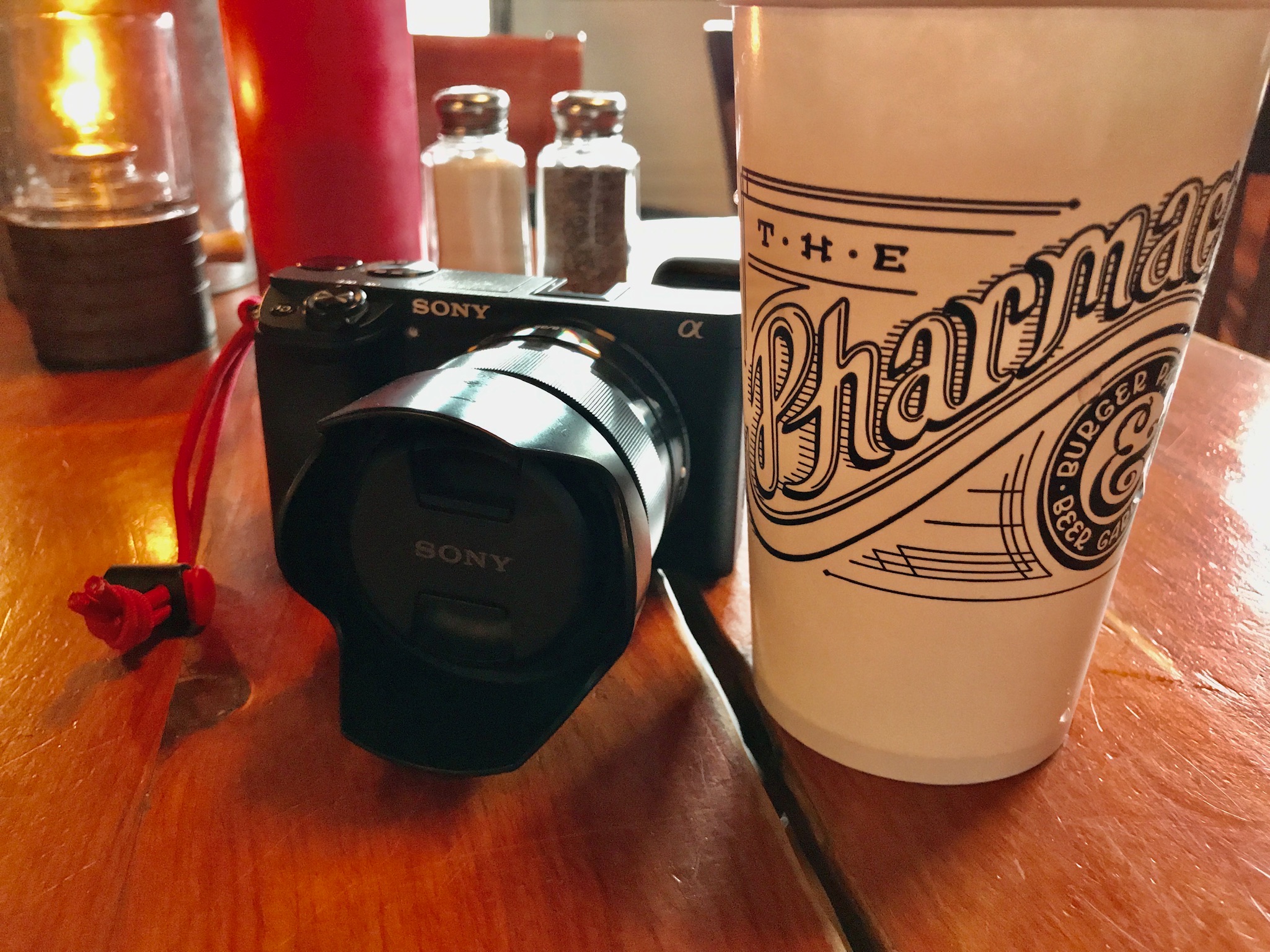 A Sony camera and a paper cup labeled "The Pharmacy" sit on a wooden table near salt and pepper shakers and a red condiment bottle.