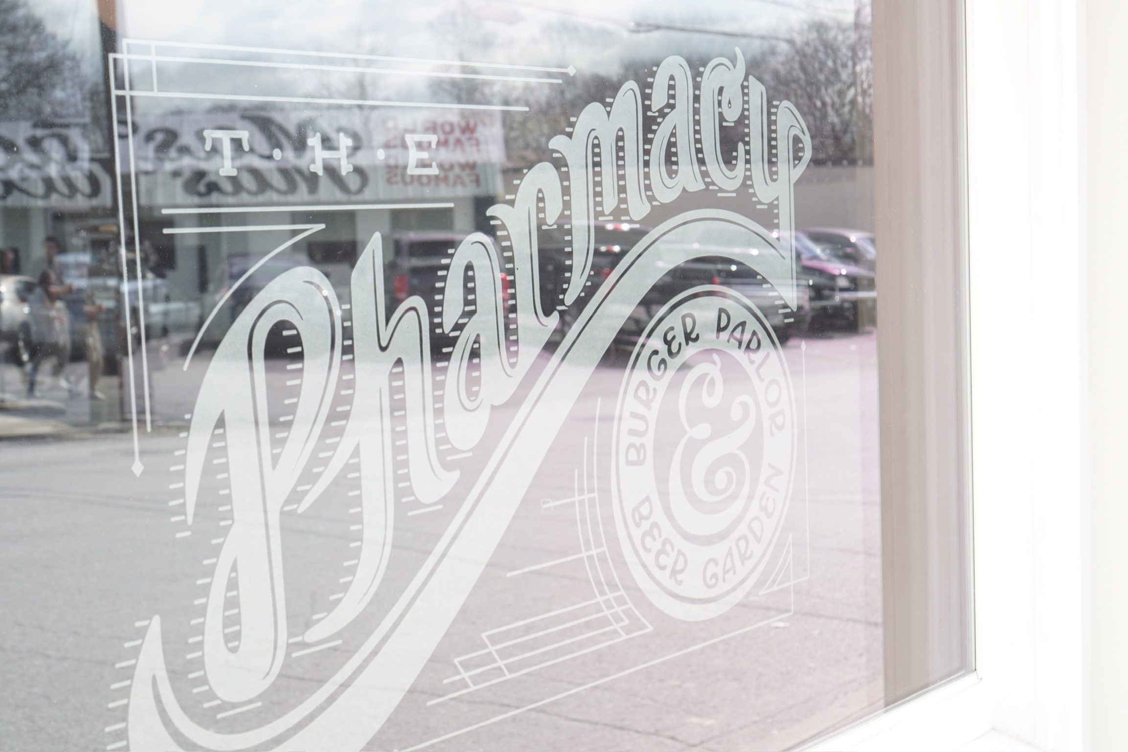 Glass window with white, ornate lettering that reads “The Pharmacy Burger Parlor & Beer Garden;” parking lot and buildings are visible in the reflection.