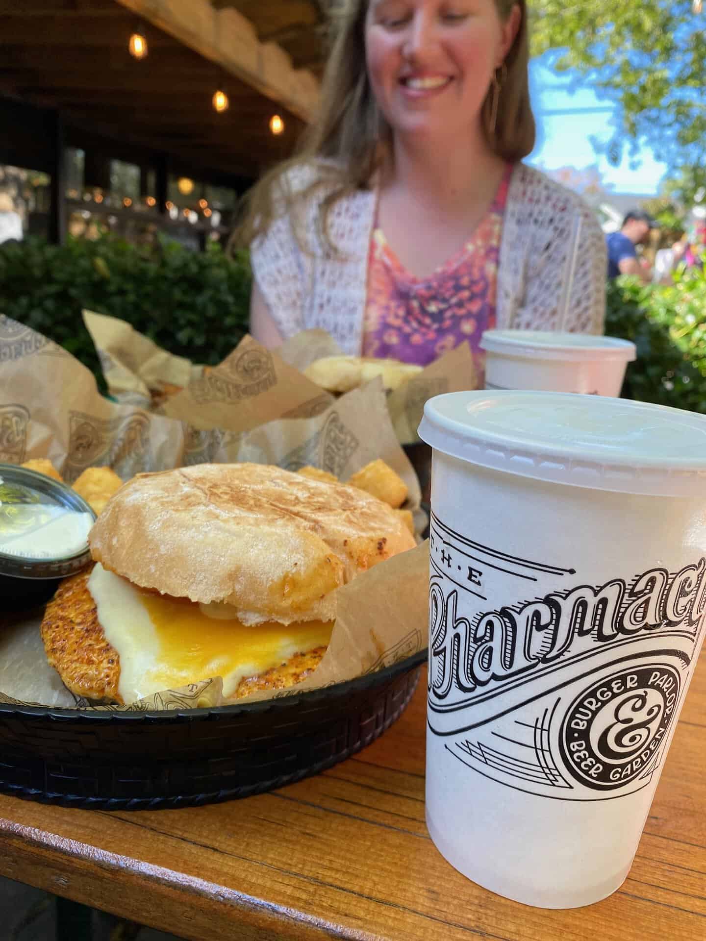 A woman sits at an outdoor table with a chicken sandwich, sauce on the side, tater tots, and a drink cup from "The Pharmacy.