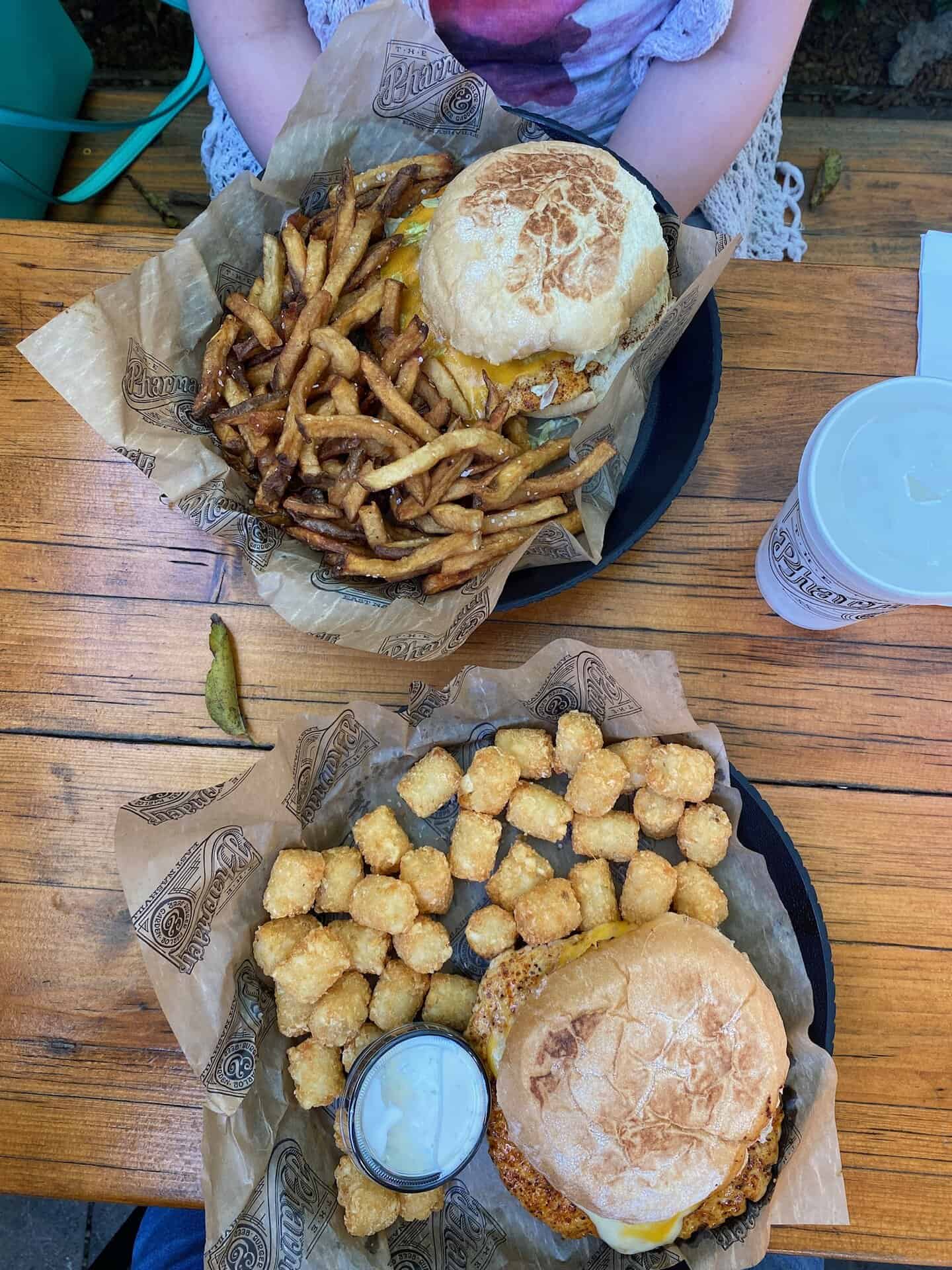 Two plates of food on a wooden table: one with a burger and fries, the other with a burger, tater tots, and a cup of sauce. A drink with a straw is visible next to the plates.