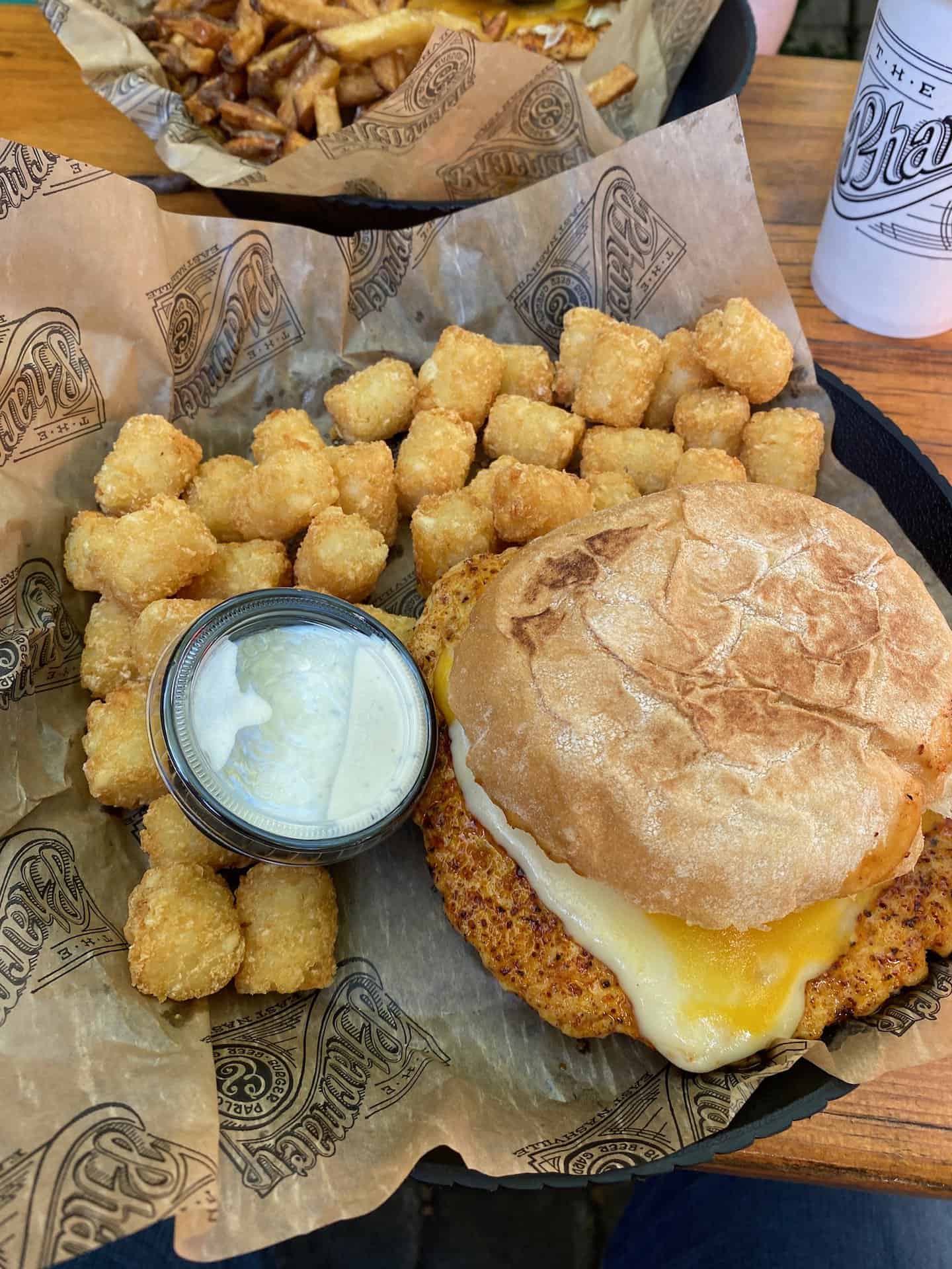 Chicken sandwich with melted cheese on a bun, served with tater tots and a cup of dipping sauce on branded paper.