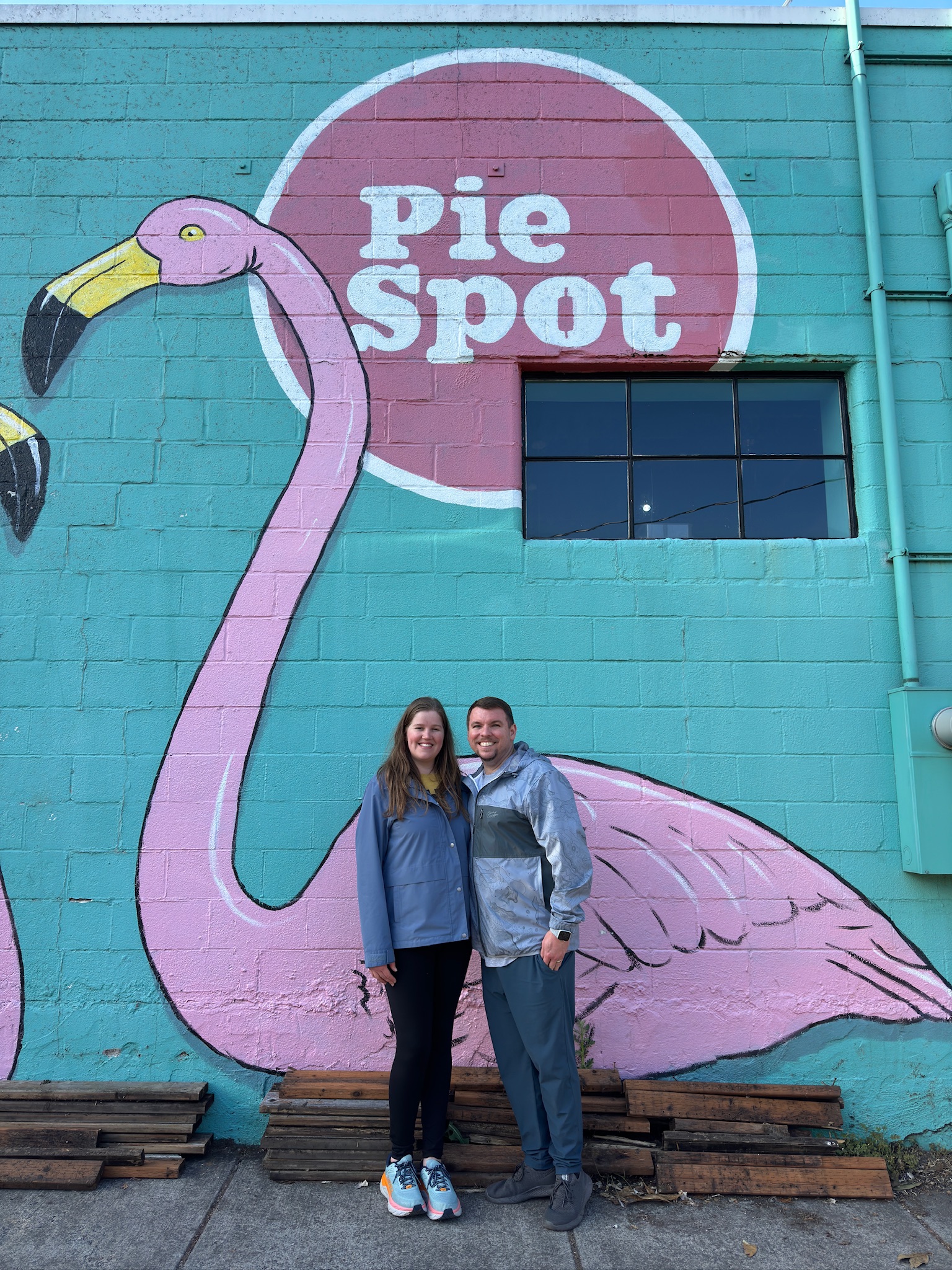 Two people stand smiling in front of a mural featuring pink flamingos and a round sign that says "Pie Spot" on a turquoise brick wall.