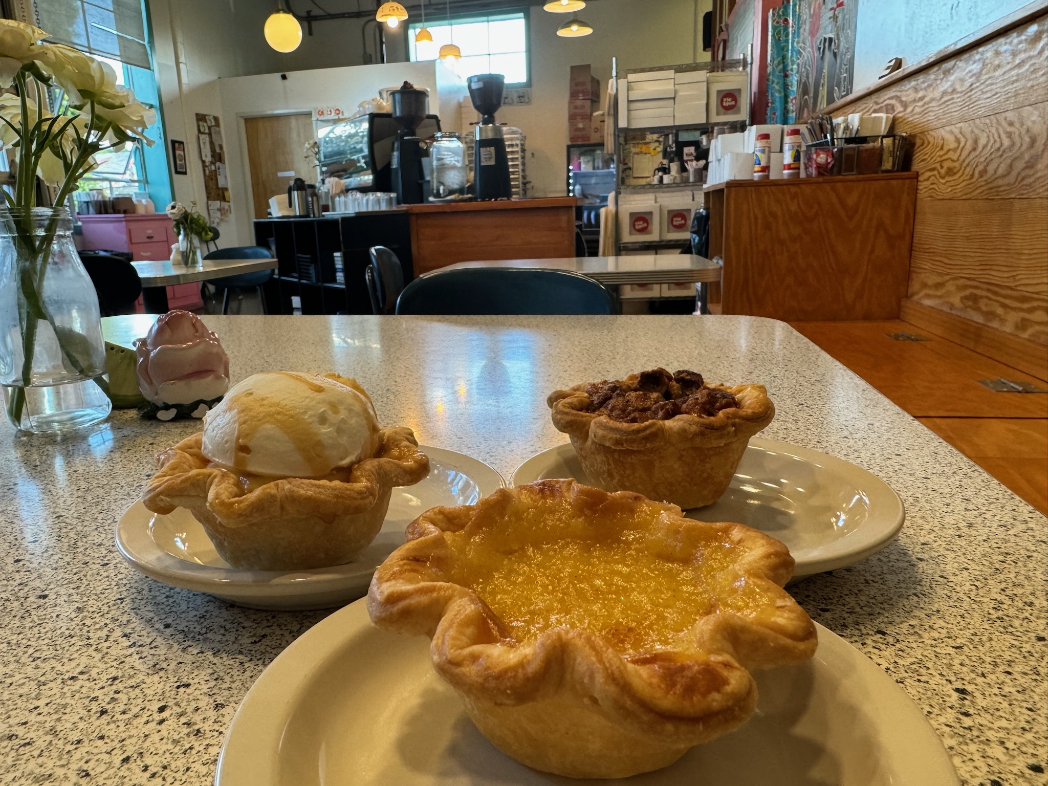 Three small pies on white plates sit on a speckled table in a cafe, with a counter and coffee machines visible in the background.