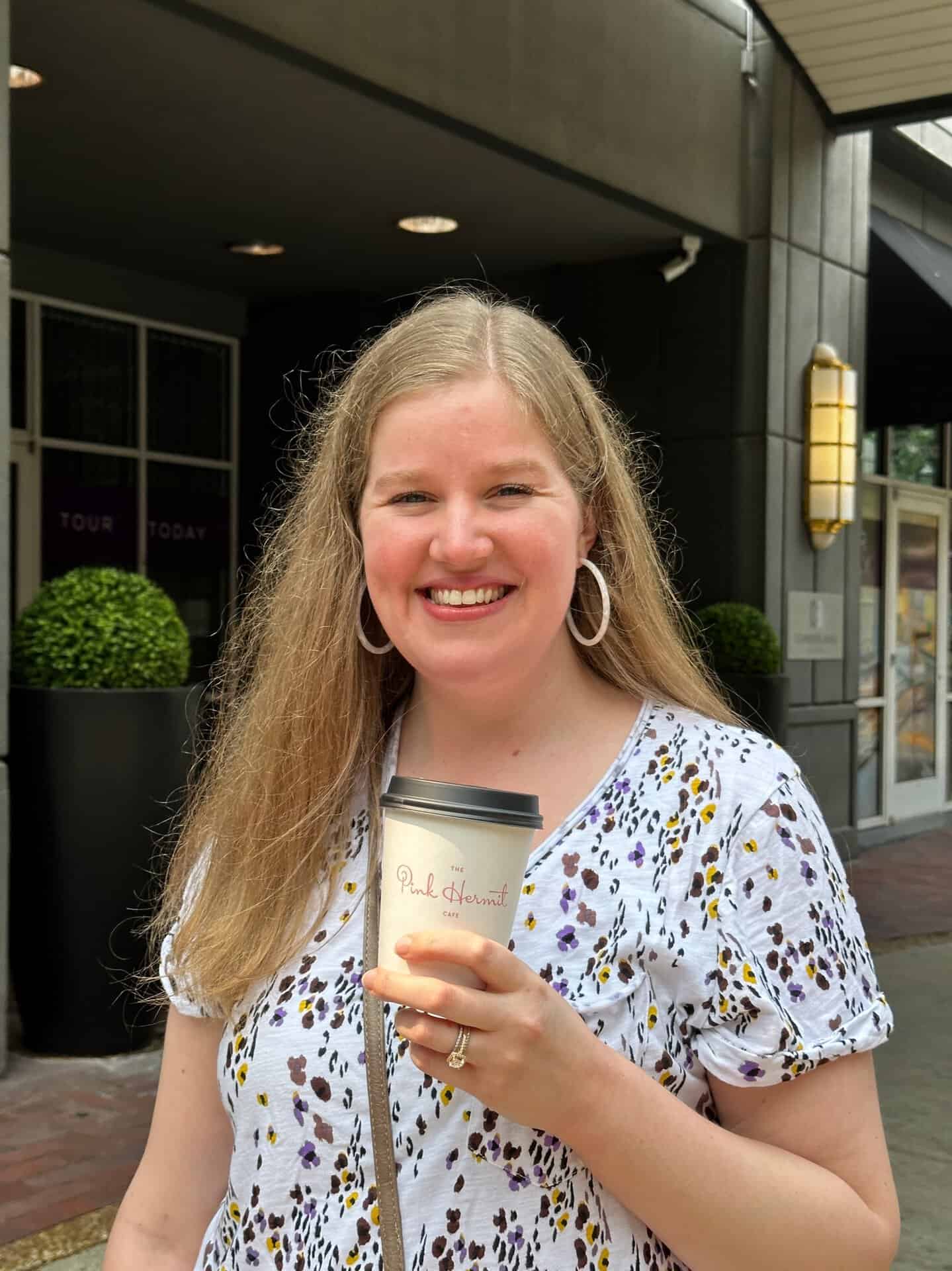 A smiling person, coffee cup in hand, stands outside a Nashville building adorned with vibrant greenery and inviting signage, perfectly capturing the city's charming essence.