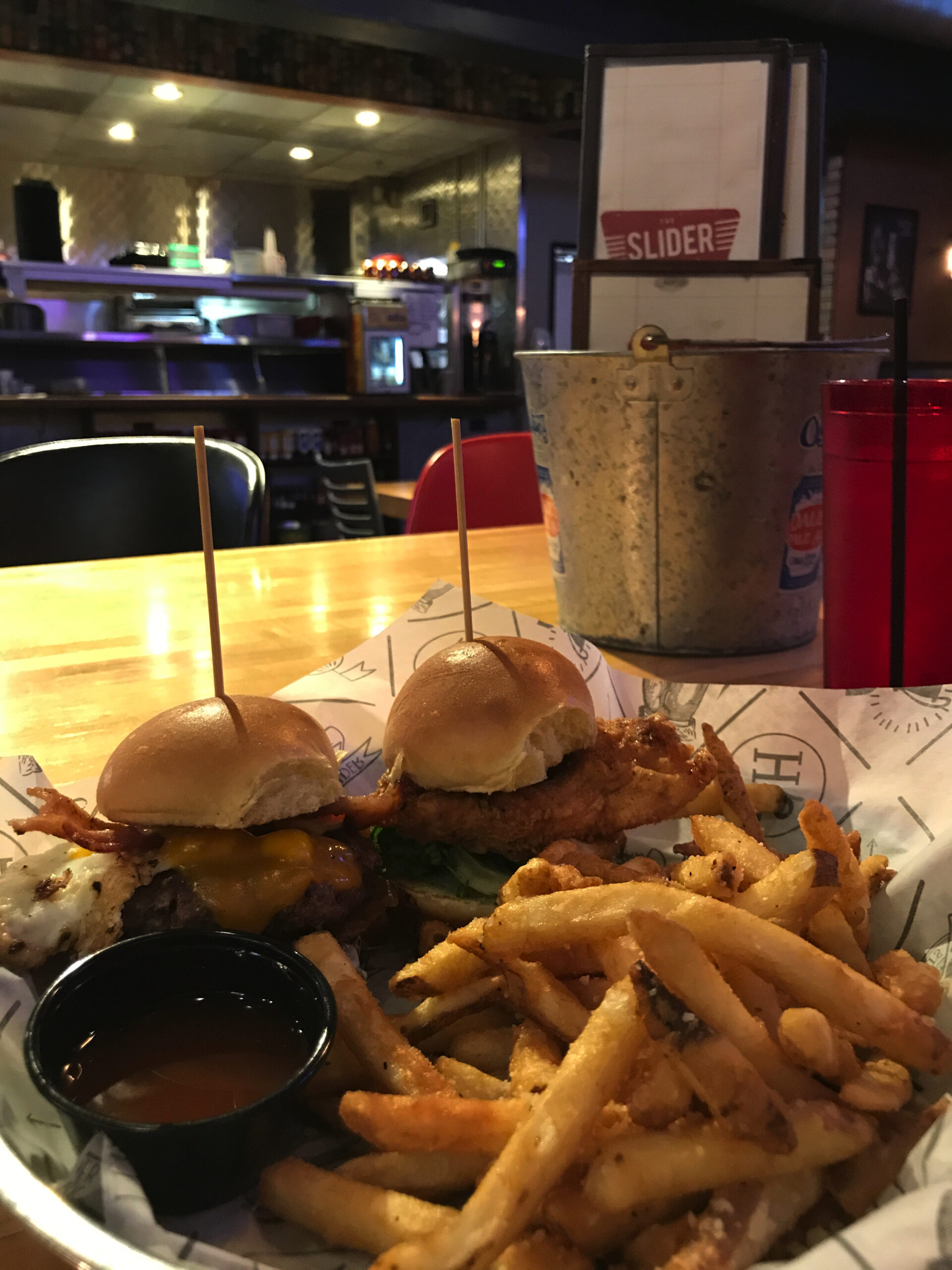 A plate with two cheeseburger sliders, fries, and a cup of sauce on a restaurant table; menus and a drink are in the background.