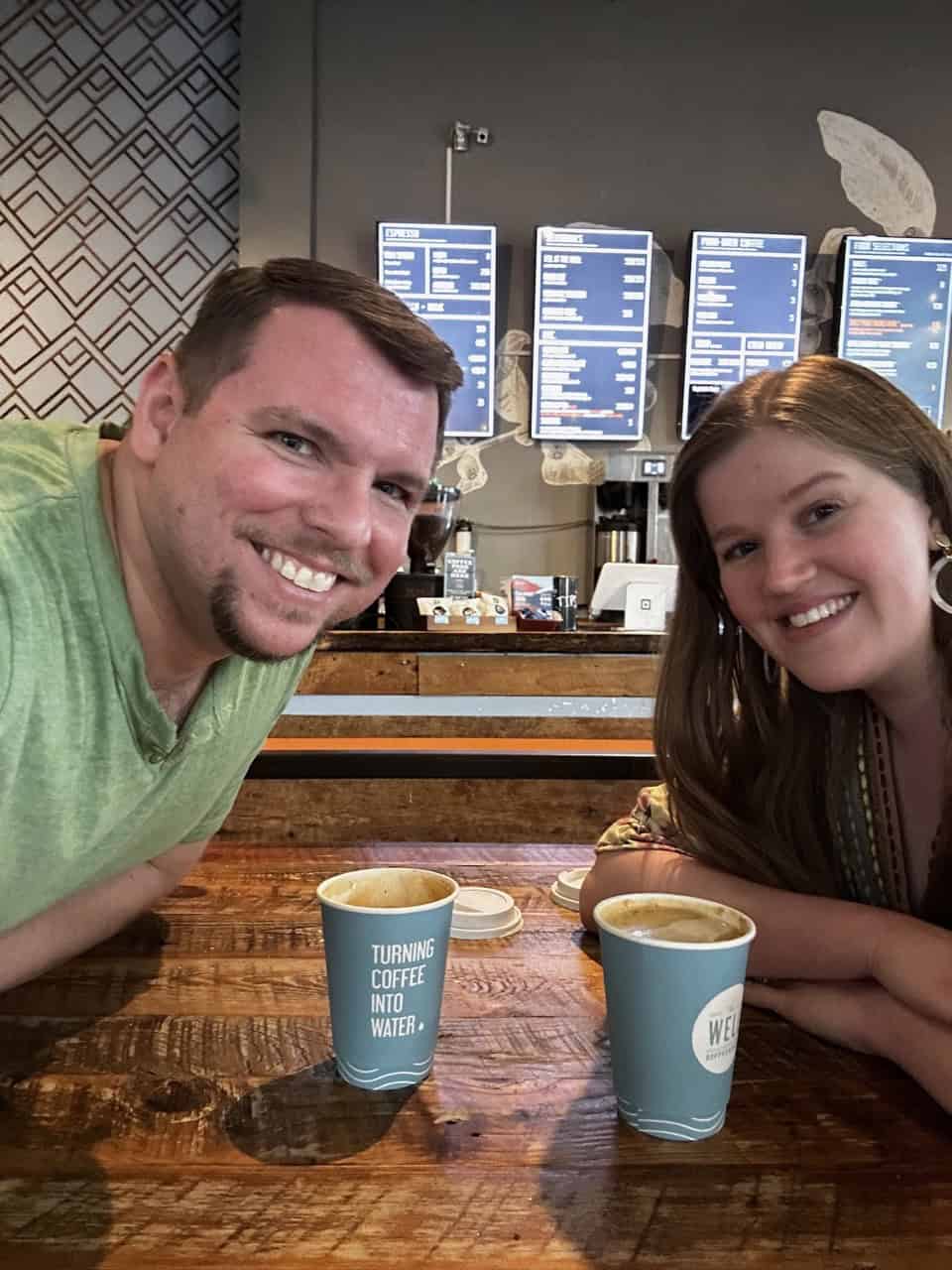 Two people smiling at a Nashville coffee shop table with two blue coffee cups. A menu board is visible in the background.