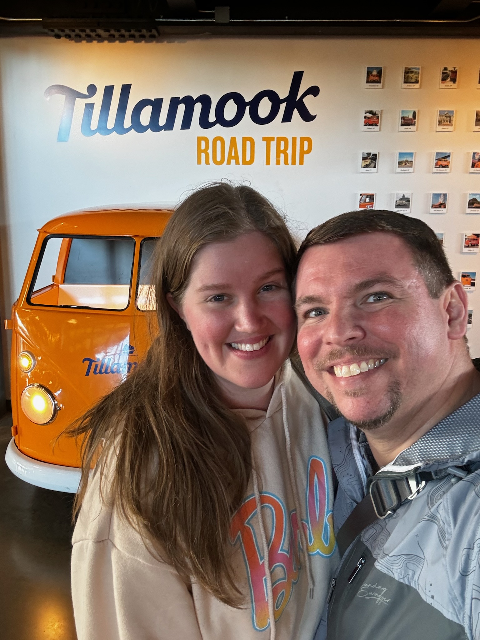 Two people smiling for a selfie in front of a small orange van and a wall sign that reads "Tillamook Road Trip.
