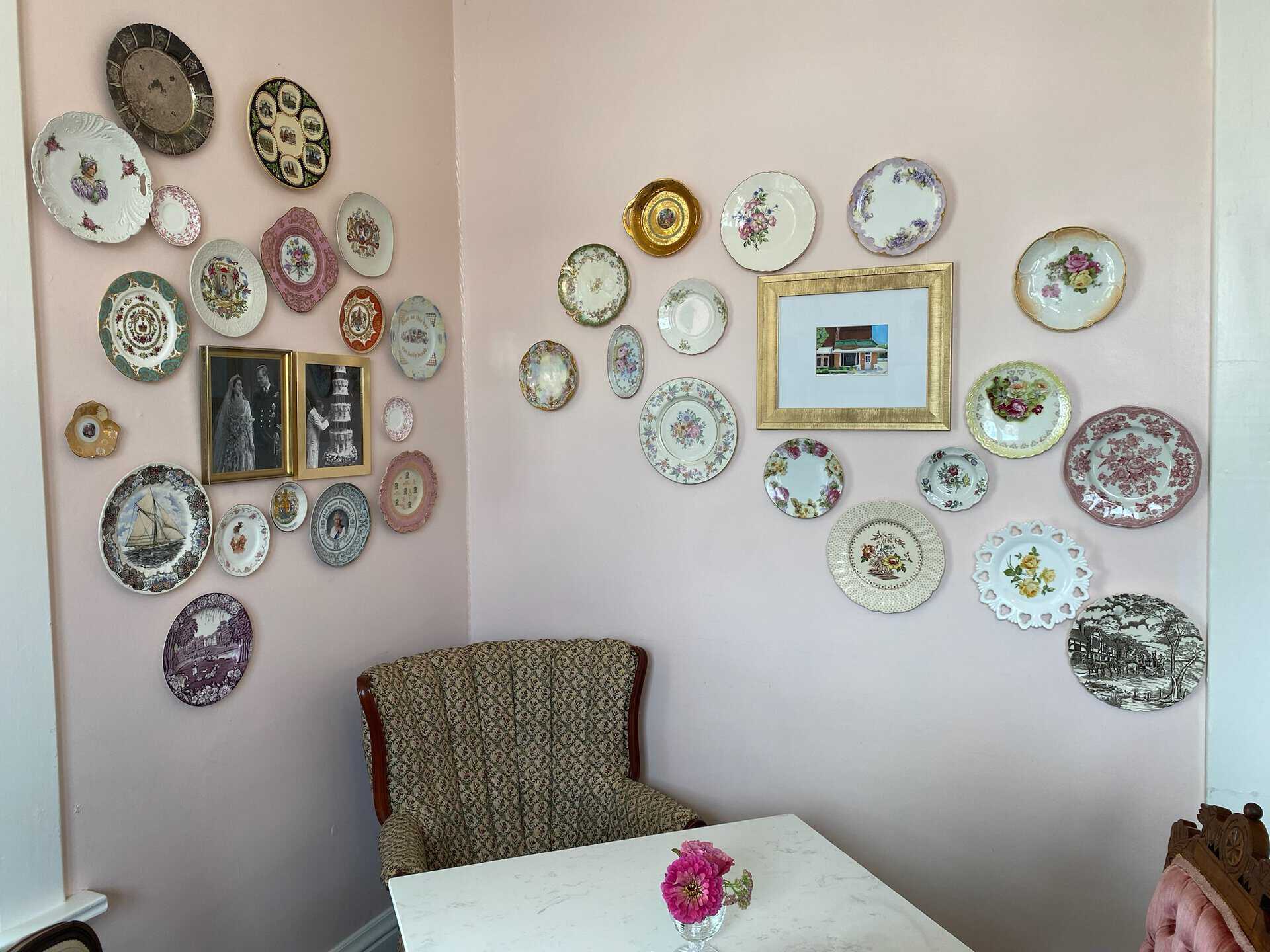 A cozy Nashville corner features a floral-patterned armchair and table. Decorative plates and framed pictures adorn the light pink walls, while a small flower arrangement adds charm to the scene.