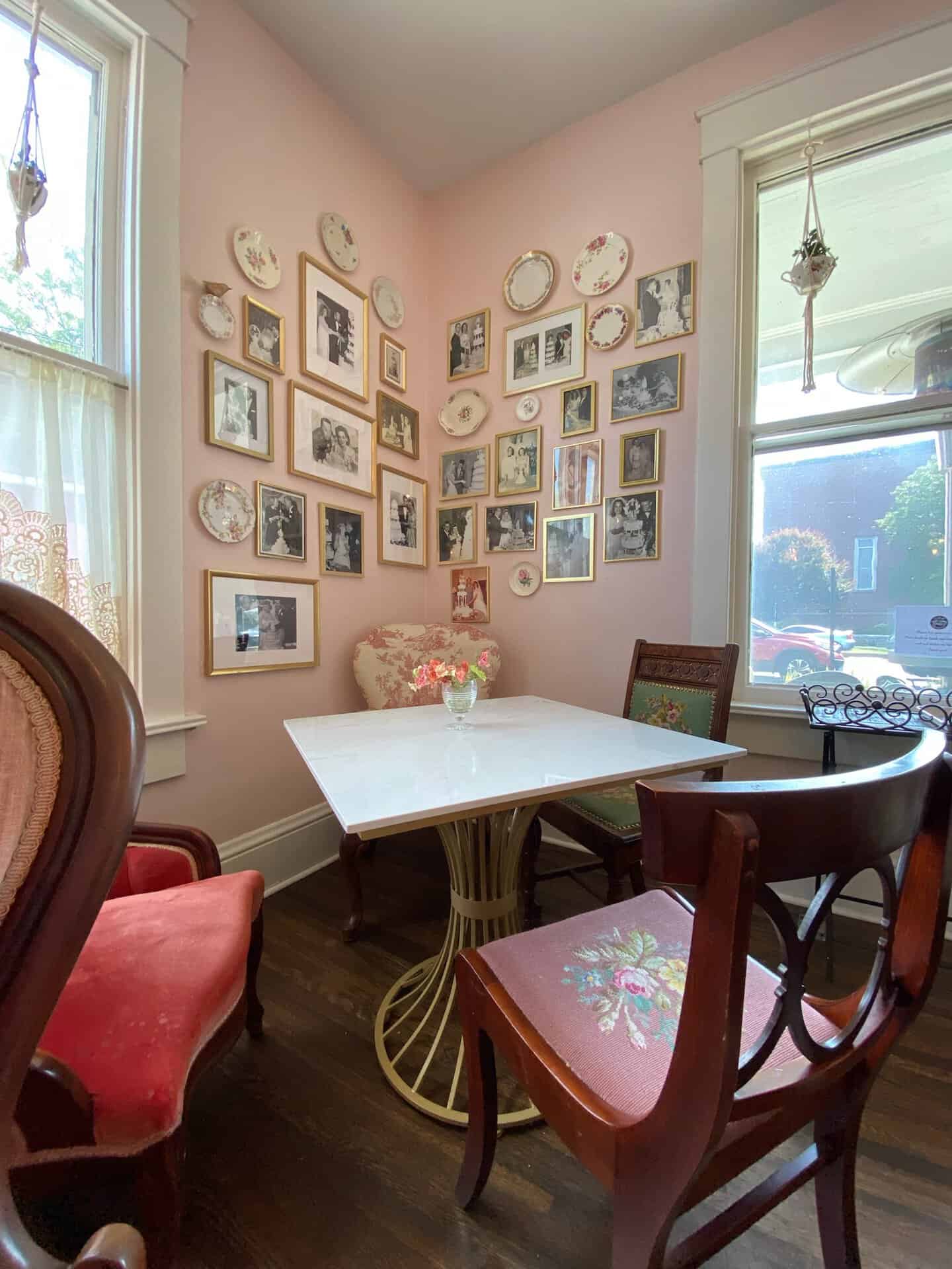 A cozy corner with a white table, surrounded by elegant chairs reminiscent of Nashville charm. Walls adorned with framed photos and decorative plates, set against a soft pink backdrop. Sunlight filters through lace-curtained windows, creating a warm Southern glow.