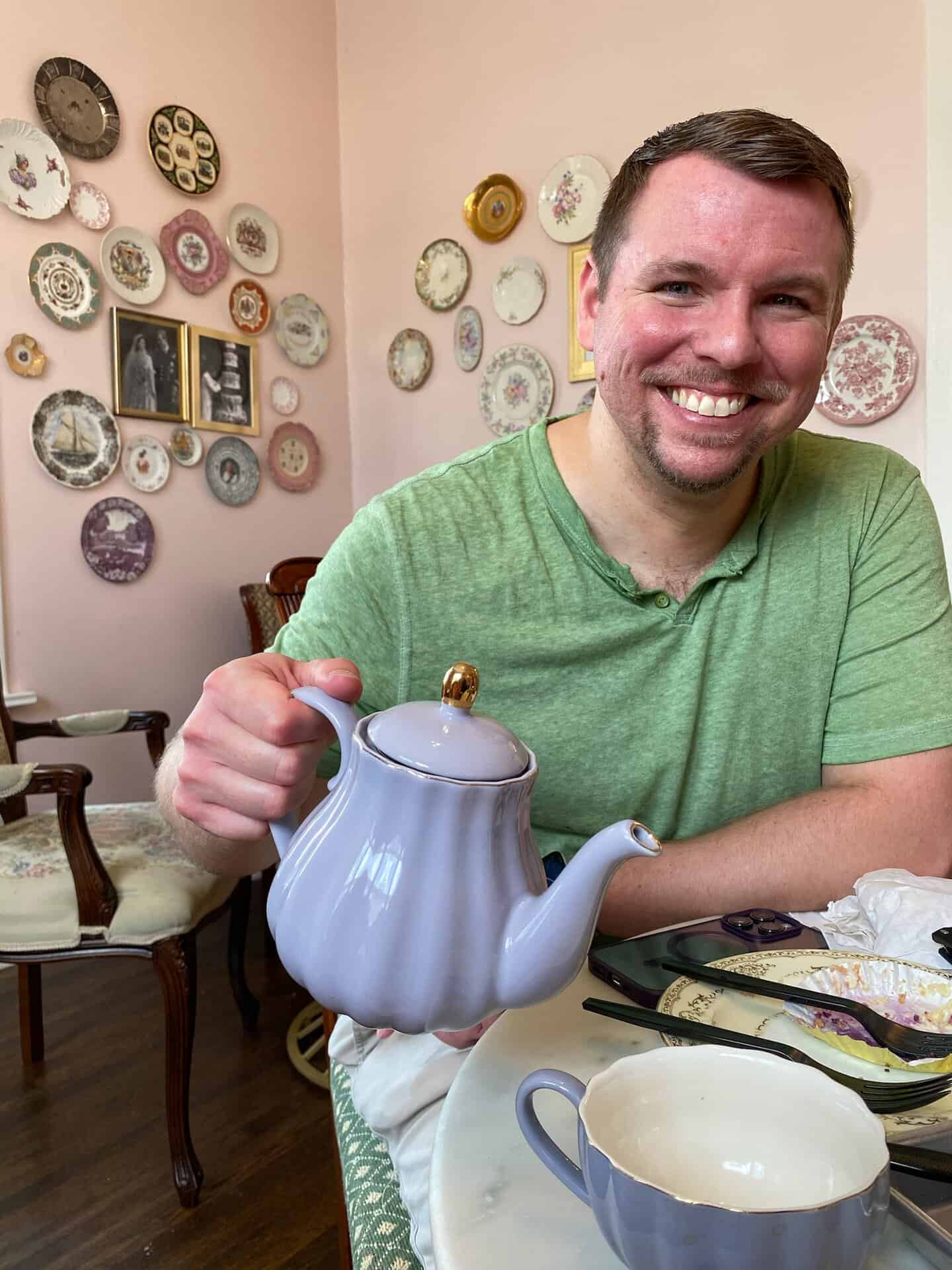 A person in a green shirt smiles warmly, holding a lavender teapot while seated at a table in Nashville, surrounded by decorative plates adorning the walls.