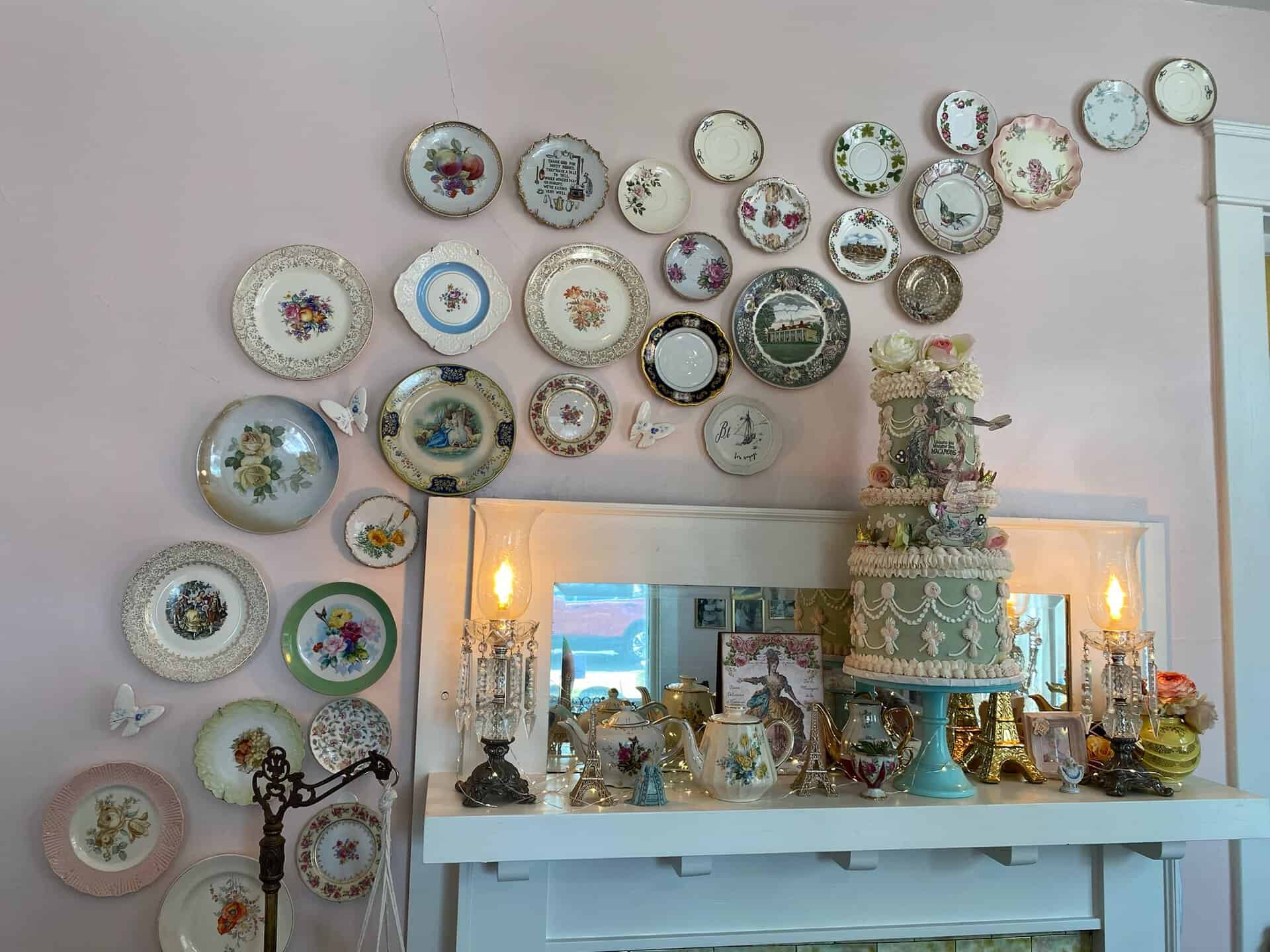 Decorative plates arranged above a mantel with various teapots, a tiered cake, and antique lamps create a charming Nashville-inspired display. Butterfly ornaments accent the decor.