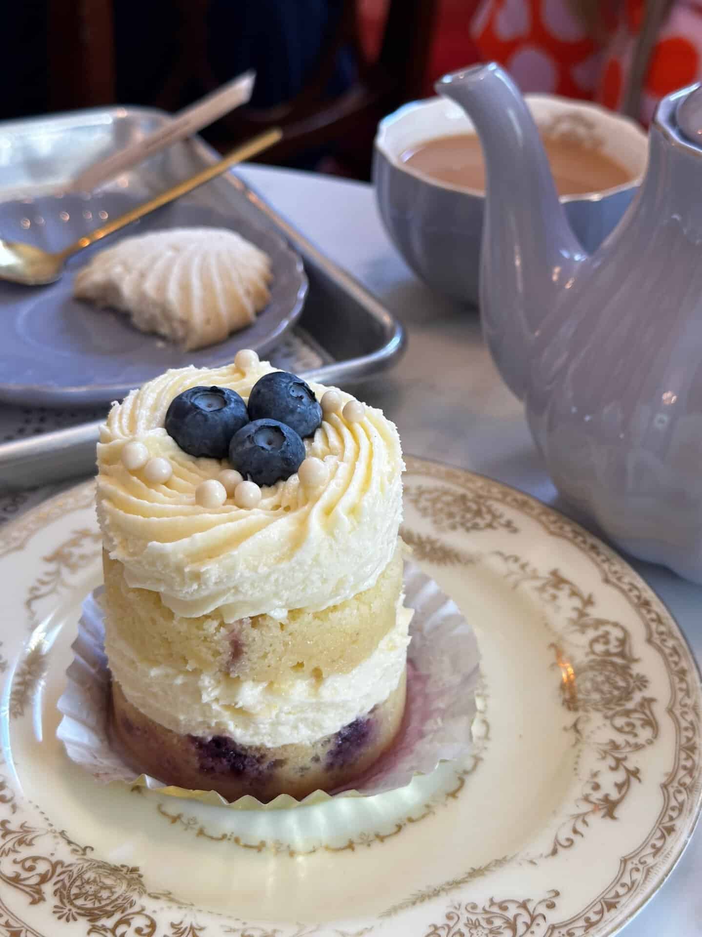 Mini cake with white frosting and blueberries on top, placed on a decorative plate, evokes a touch of Nashville charm. Next to it, a gray teapot and a partially visible cup with milk invite you for a cozy moment. A tray with a pastry rests in the background, completing this delightful scene.