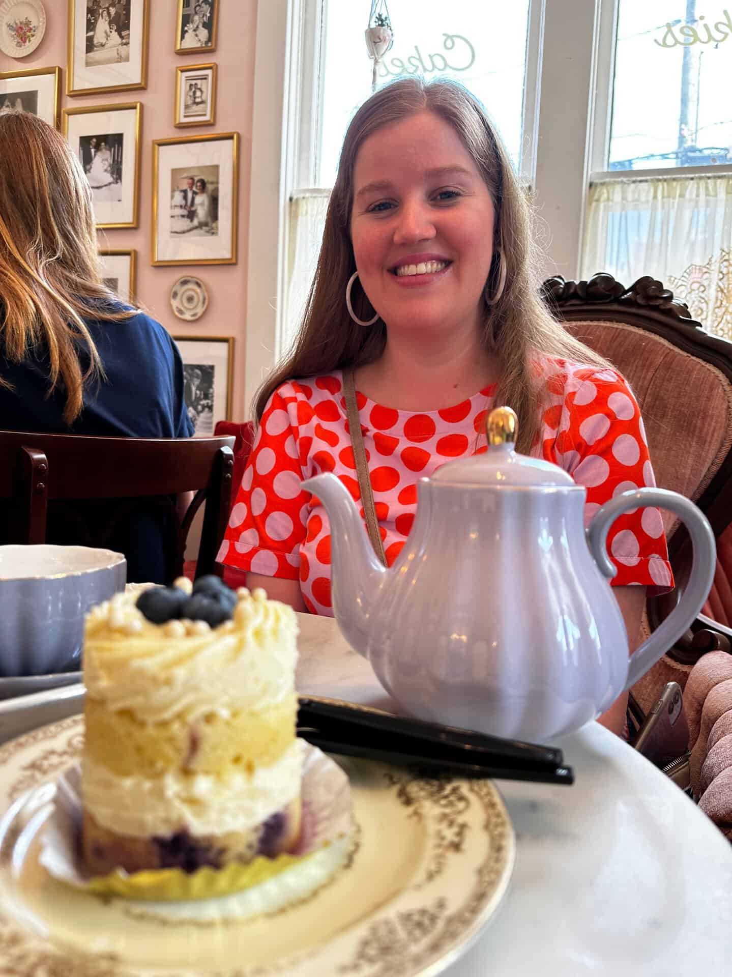 A person in a red polka dot shirt sits at a table with a teapot and dessert topped with blueberries in front of them, evoking the cozy charm of a Nashville café. Framed pictures adorn the wall behind, capturing stories in glimpses.