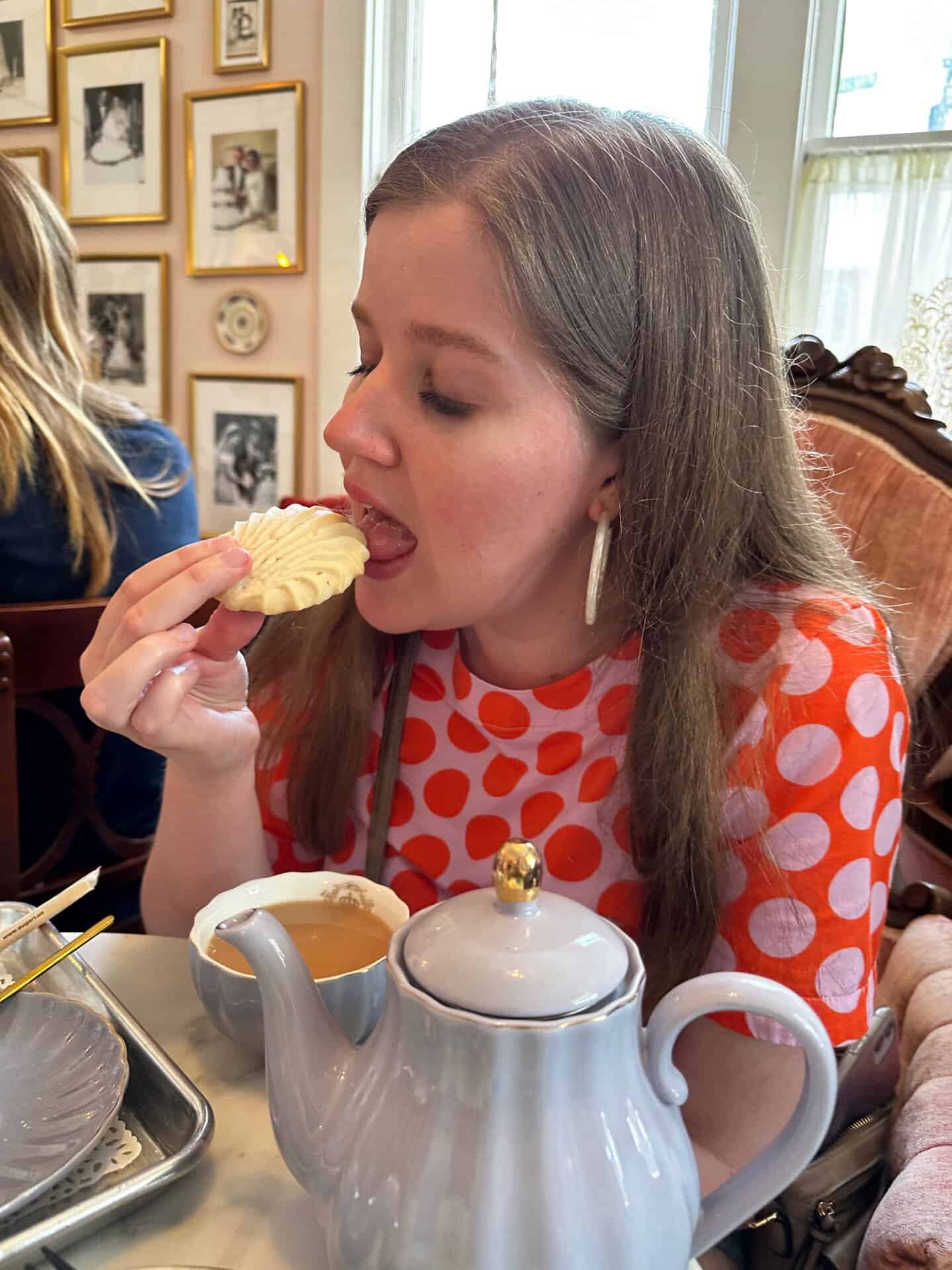In a cozy Nashville cafe, a woman in a red polka-dot shirt savors a cookie at her table. Behind her, a teapot, cup, and picture frames add to the charming ambiance.