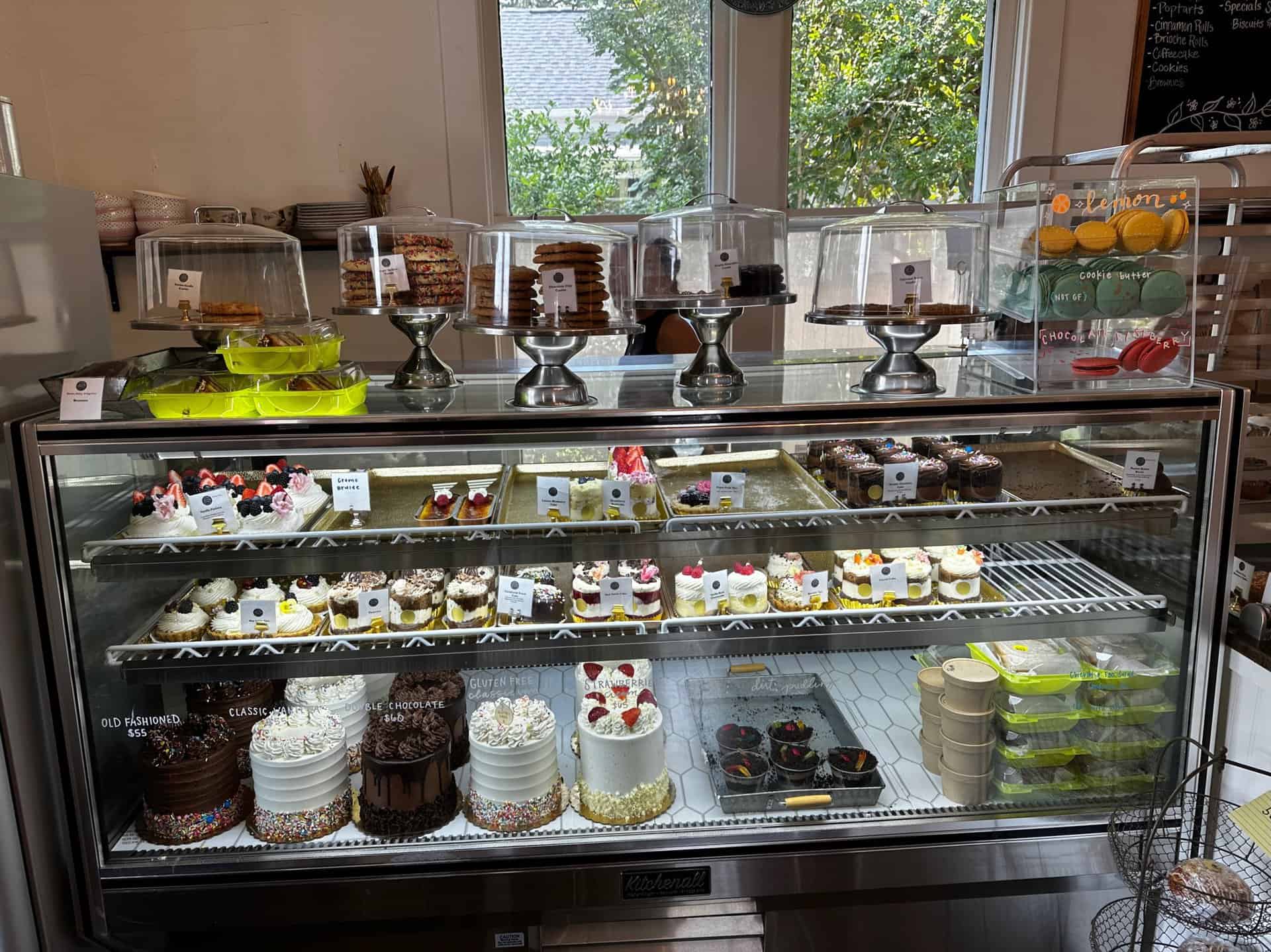 A Nashville bakery display case boasts an array of cakes, cupcakes, and pastries across multiple shelves. Delicate glass domes cover some treats, while a menu board peeks out from the right side, inviting locals and visitors alike to indulge in southern sweetness.