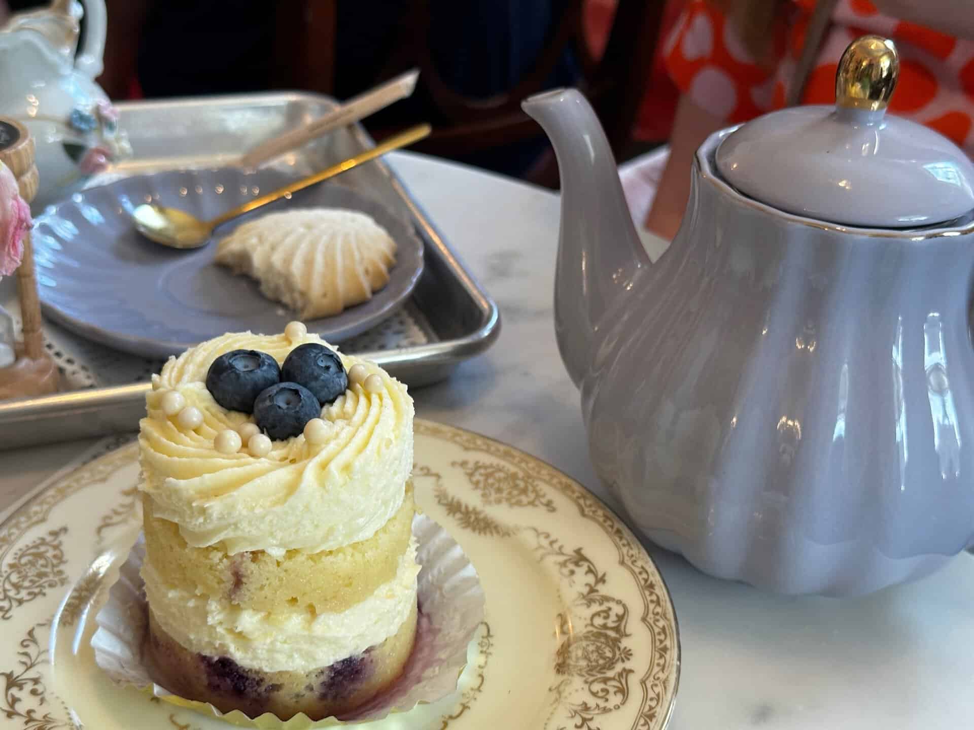 A layered cake with blueberries sits elegantly on a decorative plate, reminiscent of a charming Nashville café setting. Nearby, a ceramic white teapot and a small dessert on a tray with a spoon complete the inviting scene.