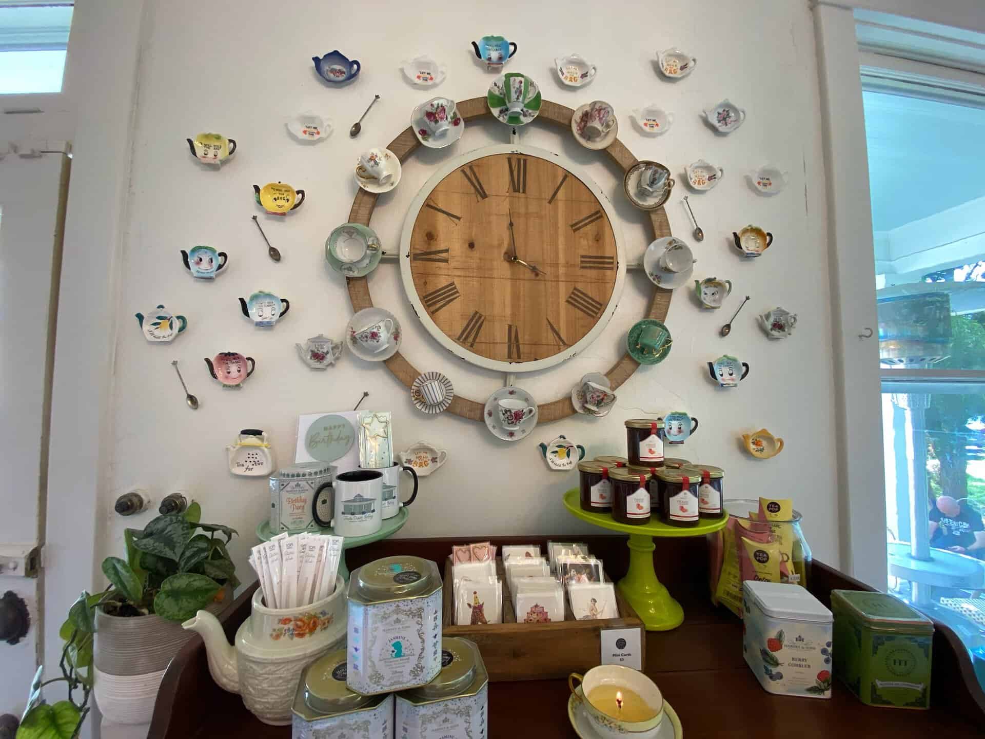 A wall clock is surrounded by decorative teacups, echoing the charm of a Nashville café. Below, a wooden surface displays various candles in jars, teapots, and potted plants. A window to the right bathes everything in natural light.