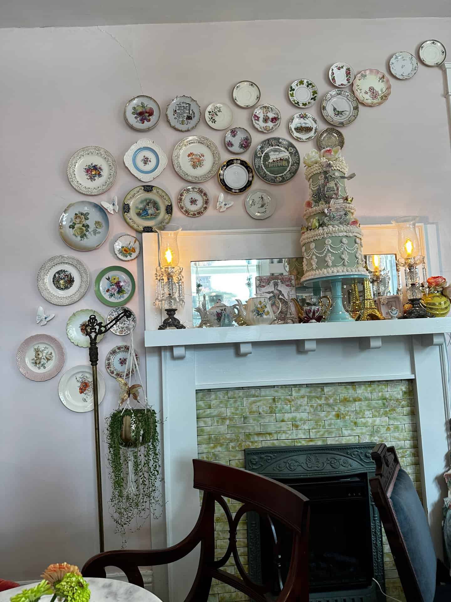 A fireplace mantel adorned with a cake, candlestick holders, and assorted decorative items evokes a touch of Nashville charm. Plates with floral and scenic designs are arranged on the wall above in a swirling pattern.