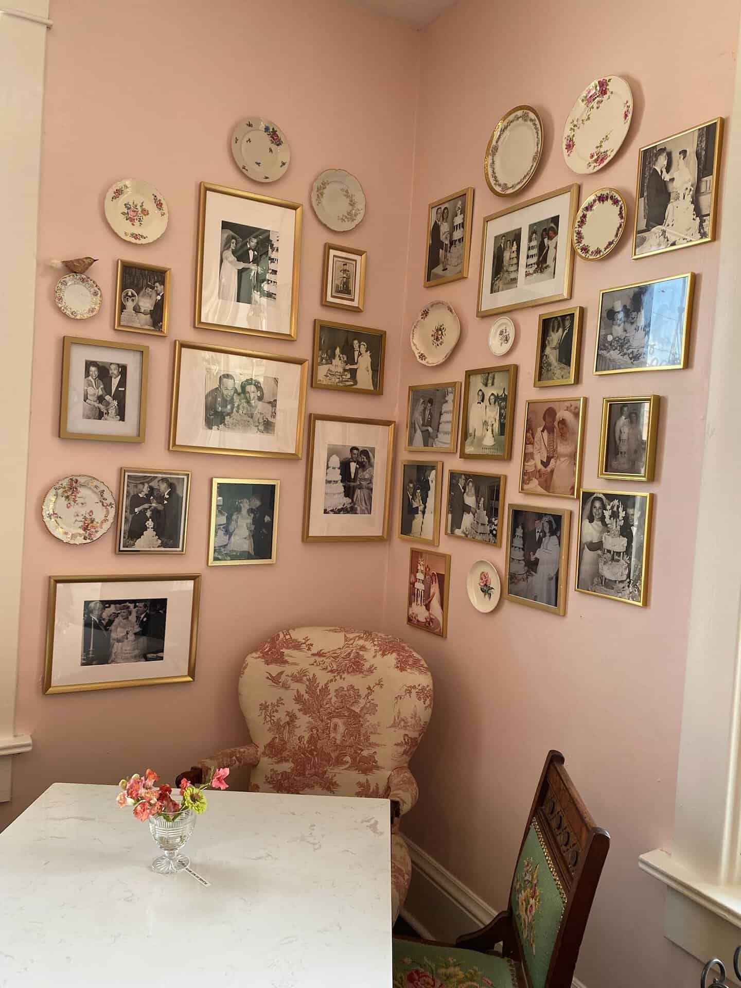 In a cozy corner of the room with pink walls, an assortment of framed photos and decorative plates tell stories. A small marble table, floral chair, and vase with flowers complete the scene, echoing the eclectic charm reminiscent of Nashville's artistic soul.
