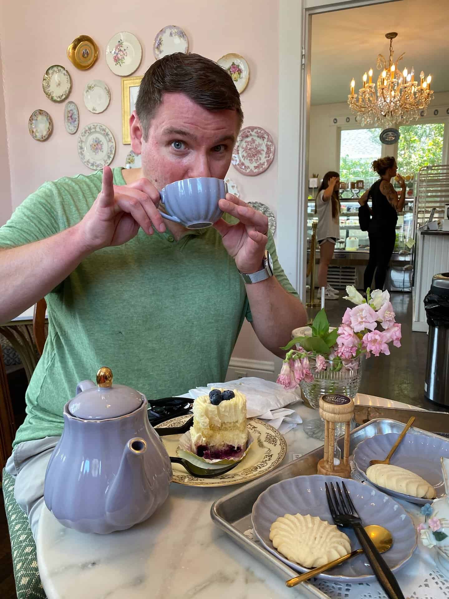 Amidst the floral decor of a Nashville café, a person in a green shirt sips tea. A slice of cake and cookies tempt from the table, alongside a charming teapot and pink flowers.