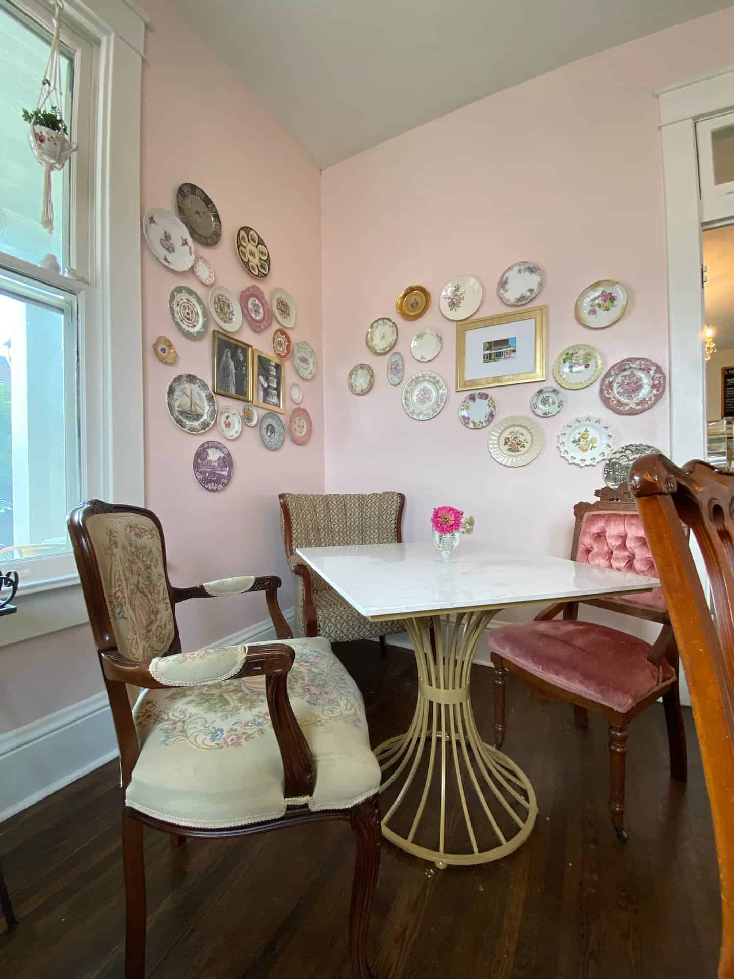 This cozy Nashville corner features a white round table, assorted chairs, and decorative plates adorning pink walls. A small vase with pink flowers adds a charming touch to the scene.