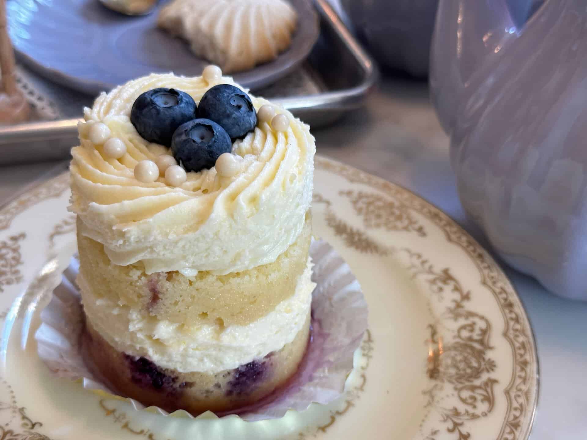 A small layered cake with white frosting, topped with blueberries and white pearls, sits on a decorative plate beside a Nashville-themed teapot.