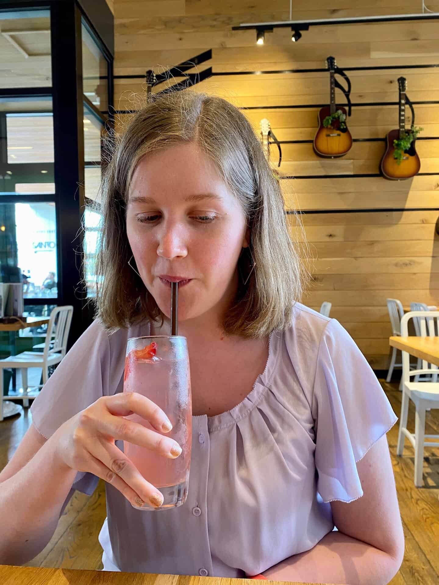A woman sips a pink beverage through a straw at a restaurant. Guitars hang on the wooden wall in the background.