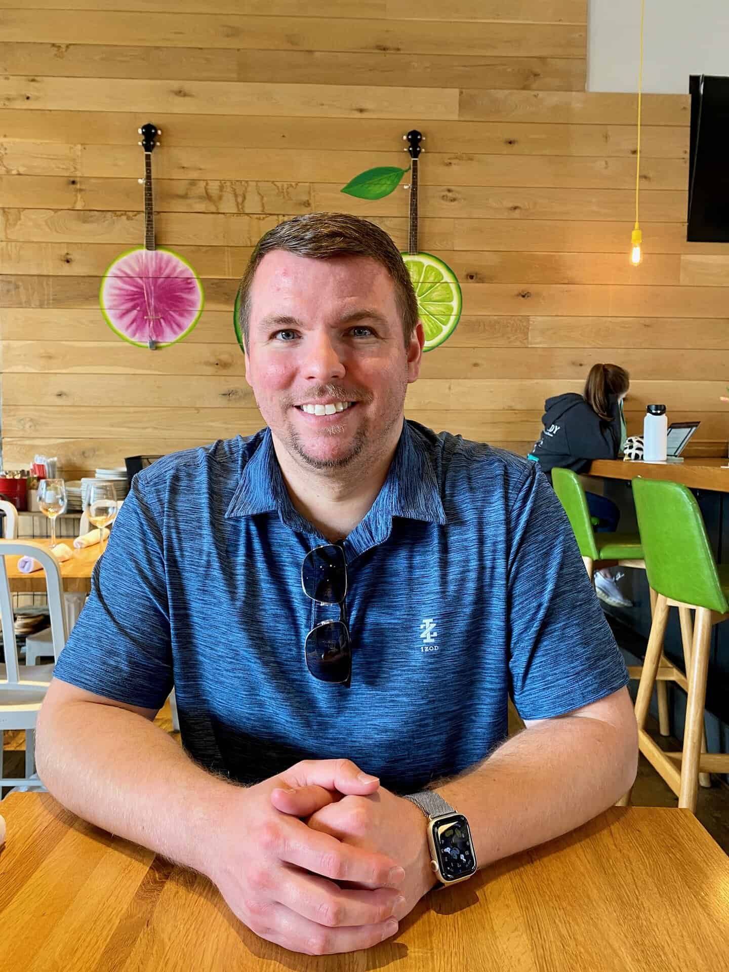 Man sitting at a wooden table in a restaurant with fruit wall decor, wearing a blue polo shirt and sunglasses on the shirt.