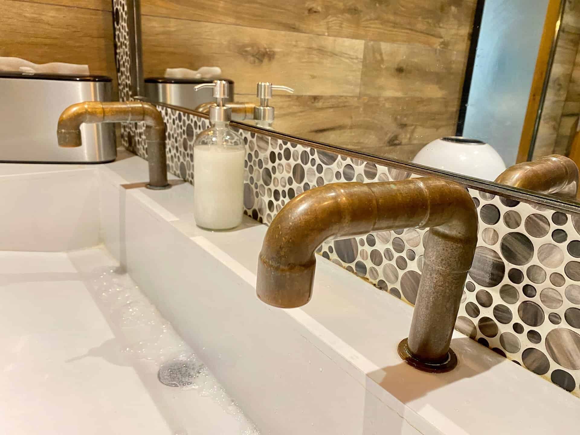 A bathroom sink with rustic copper faucets and a round pebble-patterned backsplash. A soap dispenser is positioned between the faucets, with a wooden wall in the background.