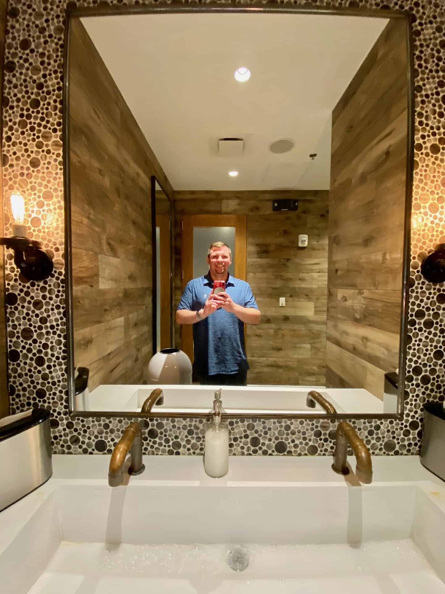 A person takes a selfie in a bathroom mirror. The room has wood-paneled walls, a white sink, and circular patterned tiles around the mirror. The person is wearing a blue shirt.