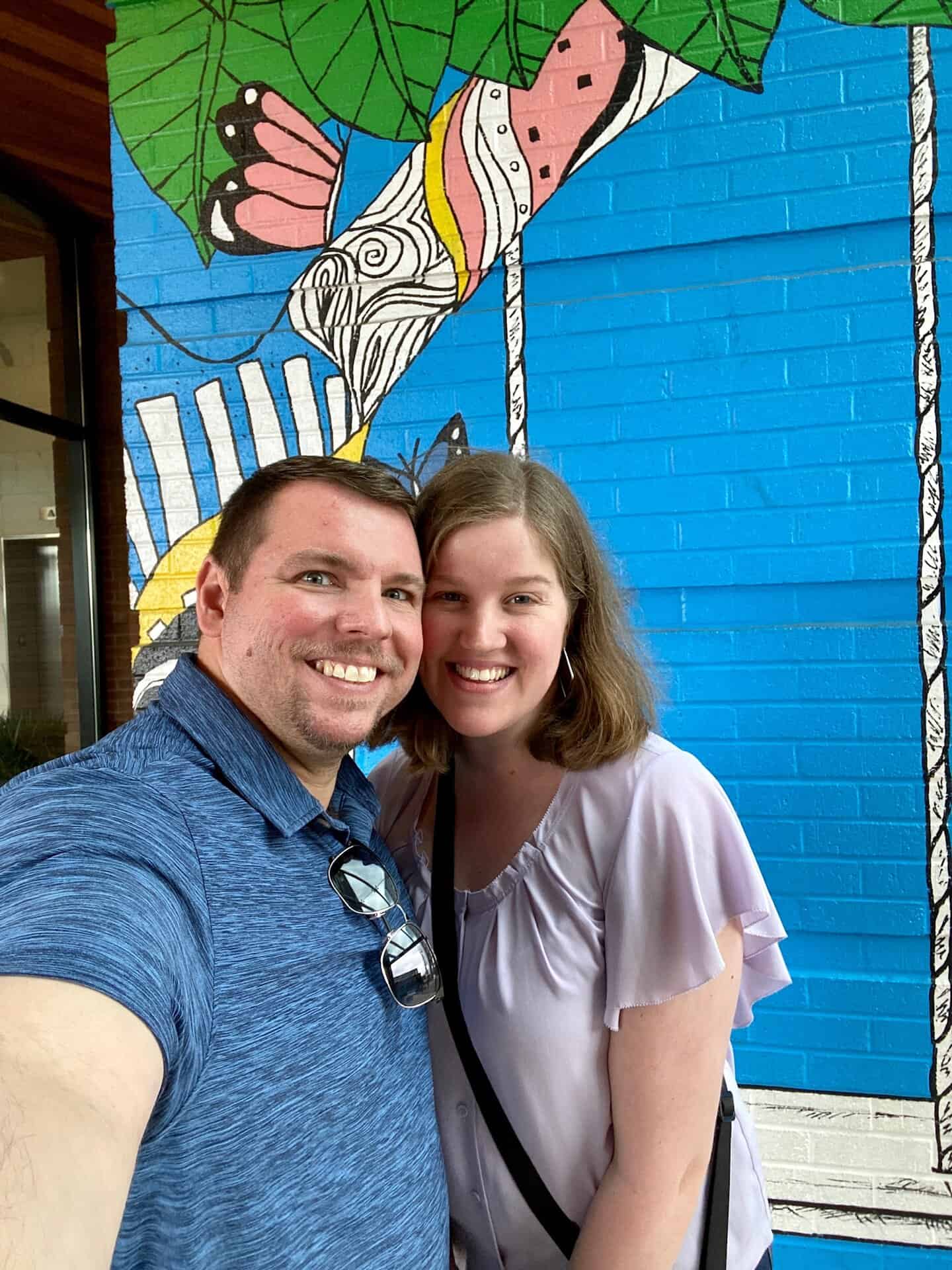 A smiling couple takes a selfie in front of a colorful mural featuring abstract shapes and leaves on a blue brick wall.