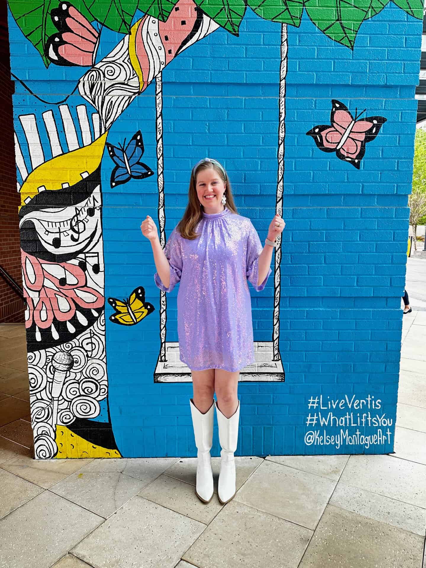 A woman in a purple dress and white boots poses in front of a vibrant Nashville mural featuring butterflies and a swing.