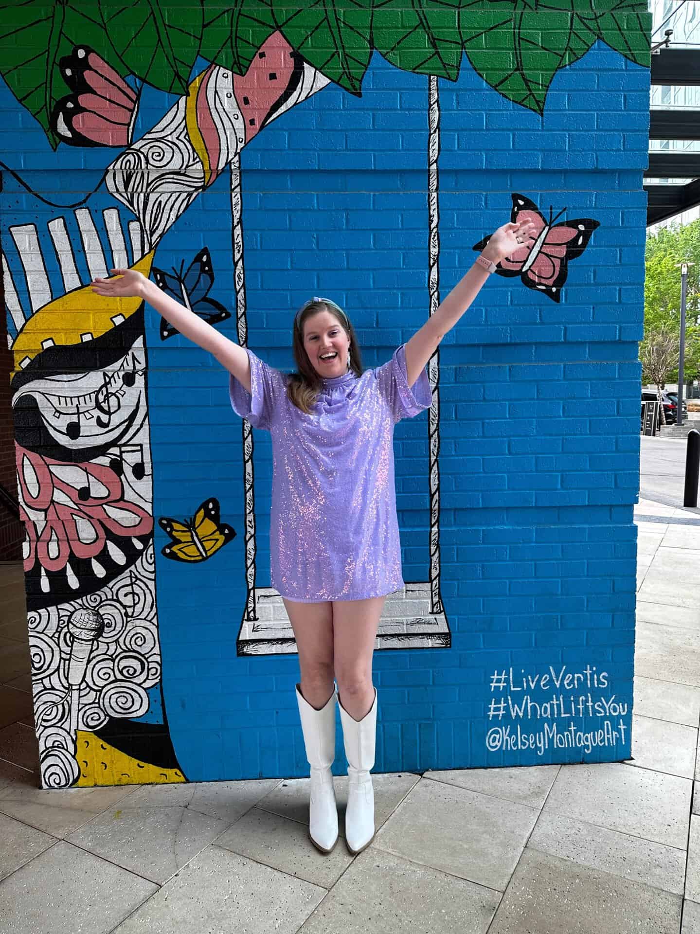 A person in a purple dress and white boots stands with arms raised, embracing the vibrant spirit of Nashville in front of a colorful mural adorned with butterflies, a swing, and lush greenery.