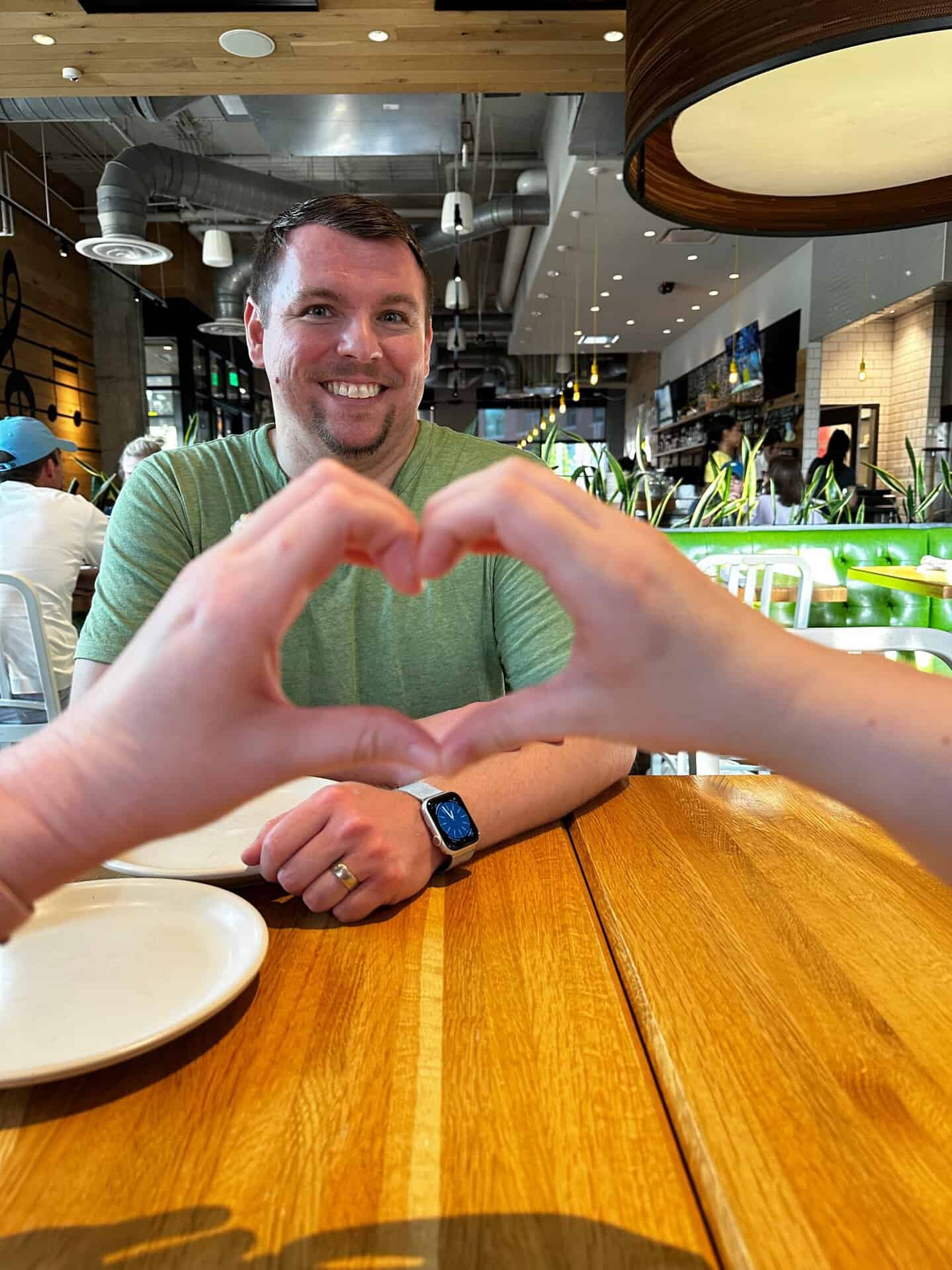 Two hands form a heart shape in the foreground, framing a smiling man seated at a wooden table in a modern Nashville restaurant.