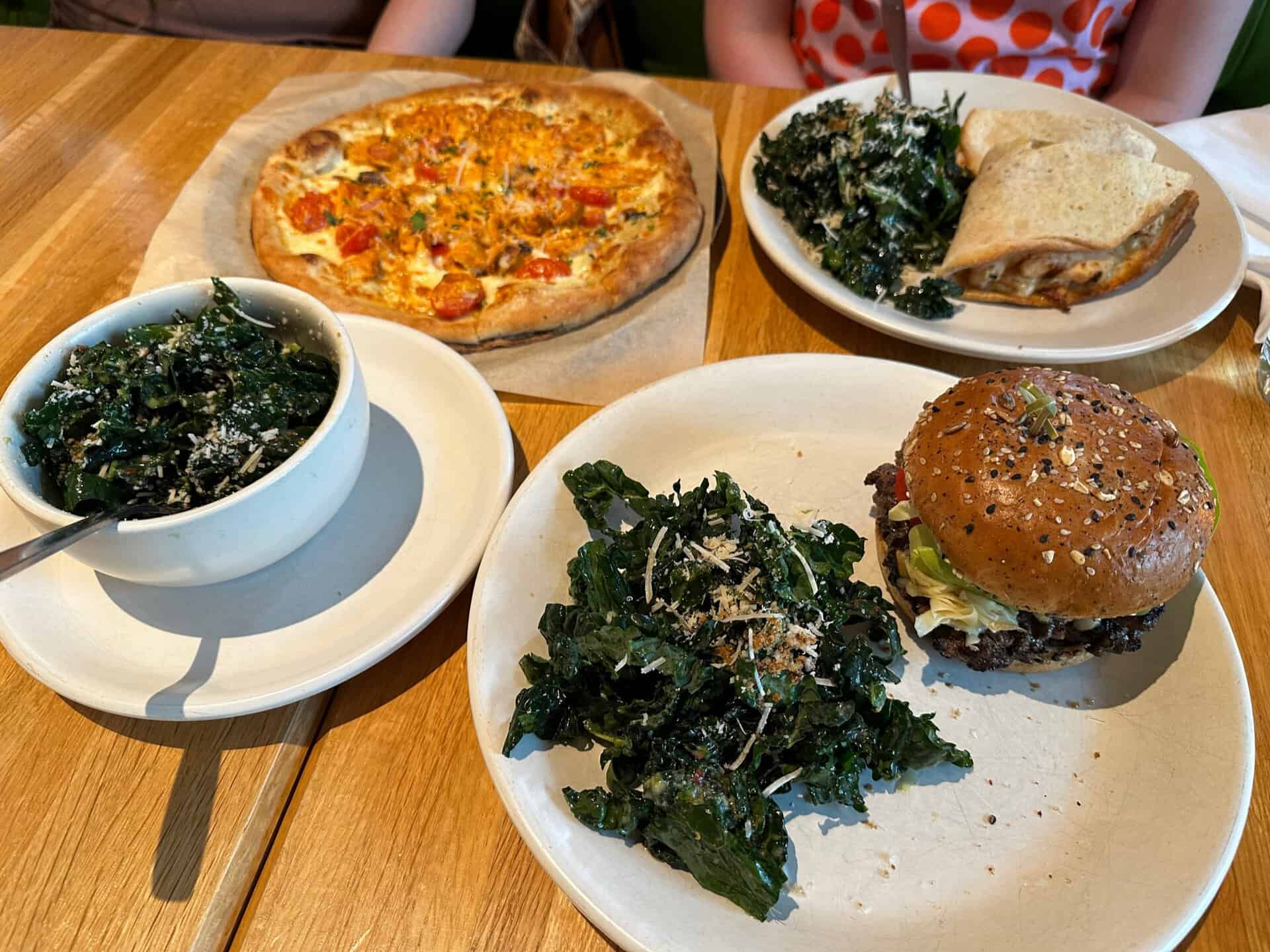 Plates of food, including a Nashville hot chicken pizza, a burger with a bun, a sandwich, and bowls of leafy green salad, are beautifully arranged on a wooden table.